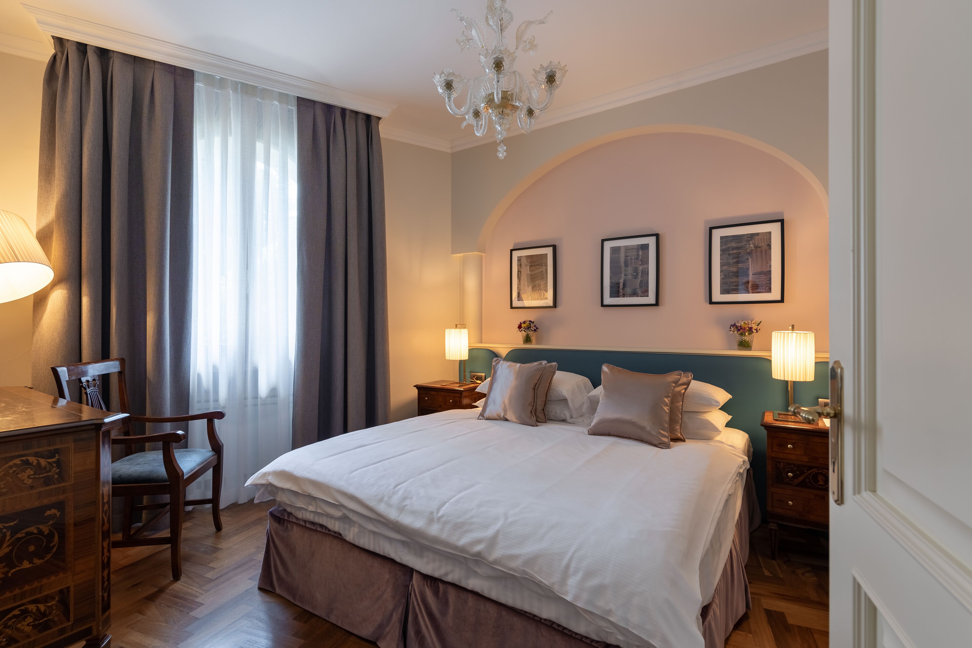 Hotelkamer met een groot bed, een bureau, een stoel en een kroonluchter. Er staan twee nachtkastjes met lampen bij en aan de muur hangen afbeeldingen in lijsten.
