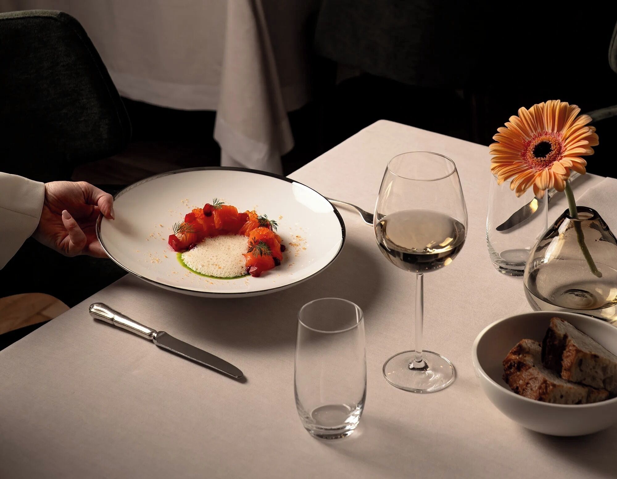 A dining table with a plate of food, a wine glass, a flower vase, and a knife.