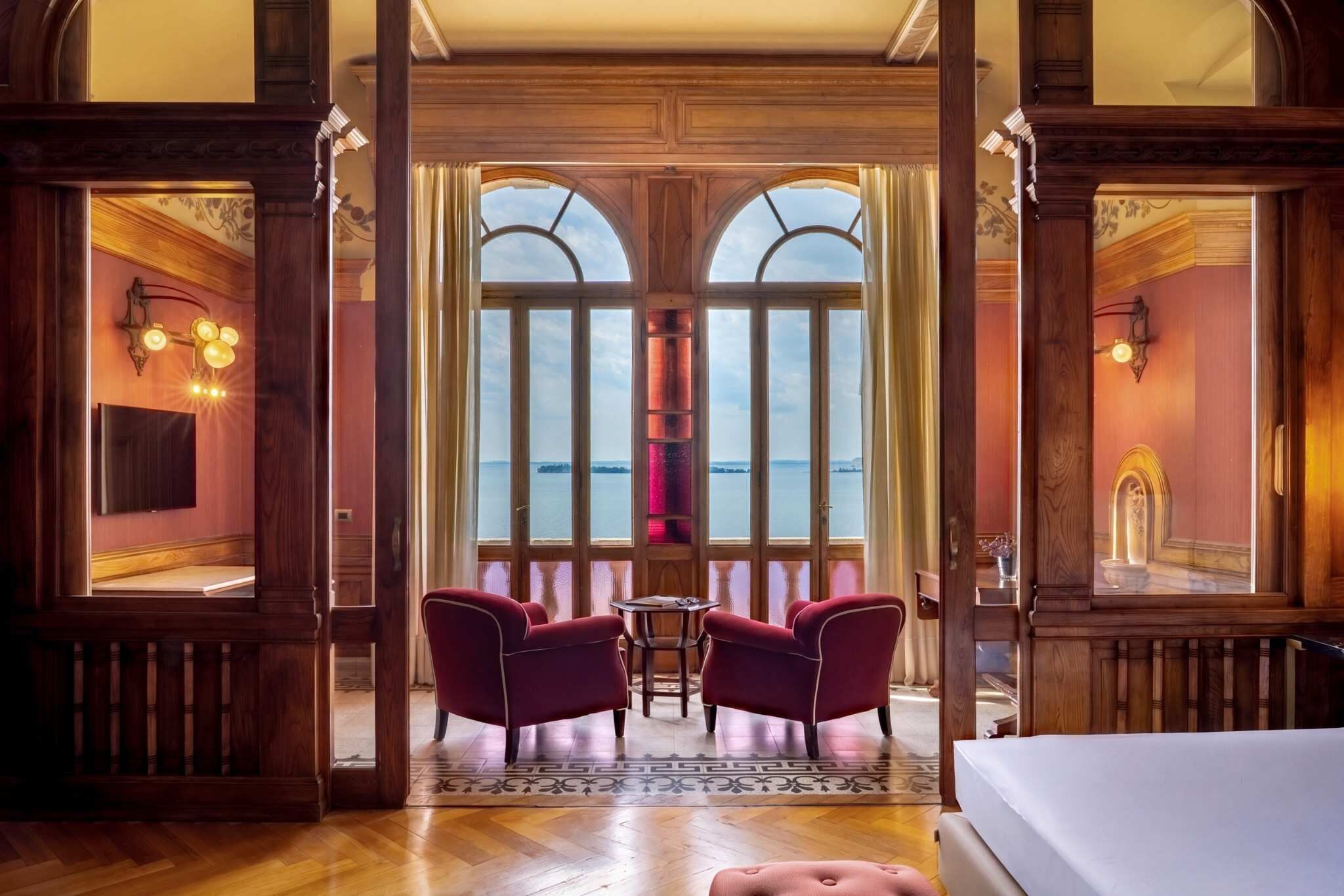 Una habitación con un ventanal grande, dos sillas rojas, una mesita y una cama.