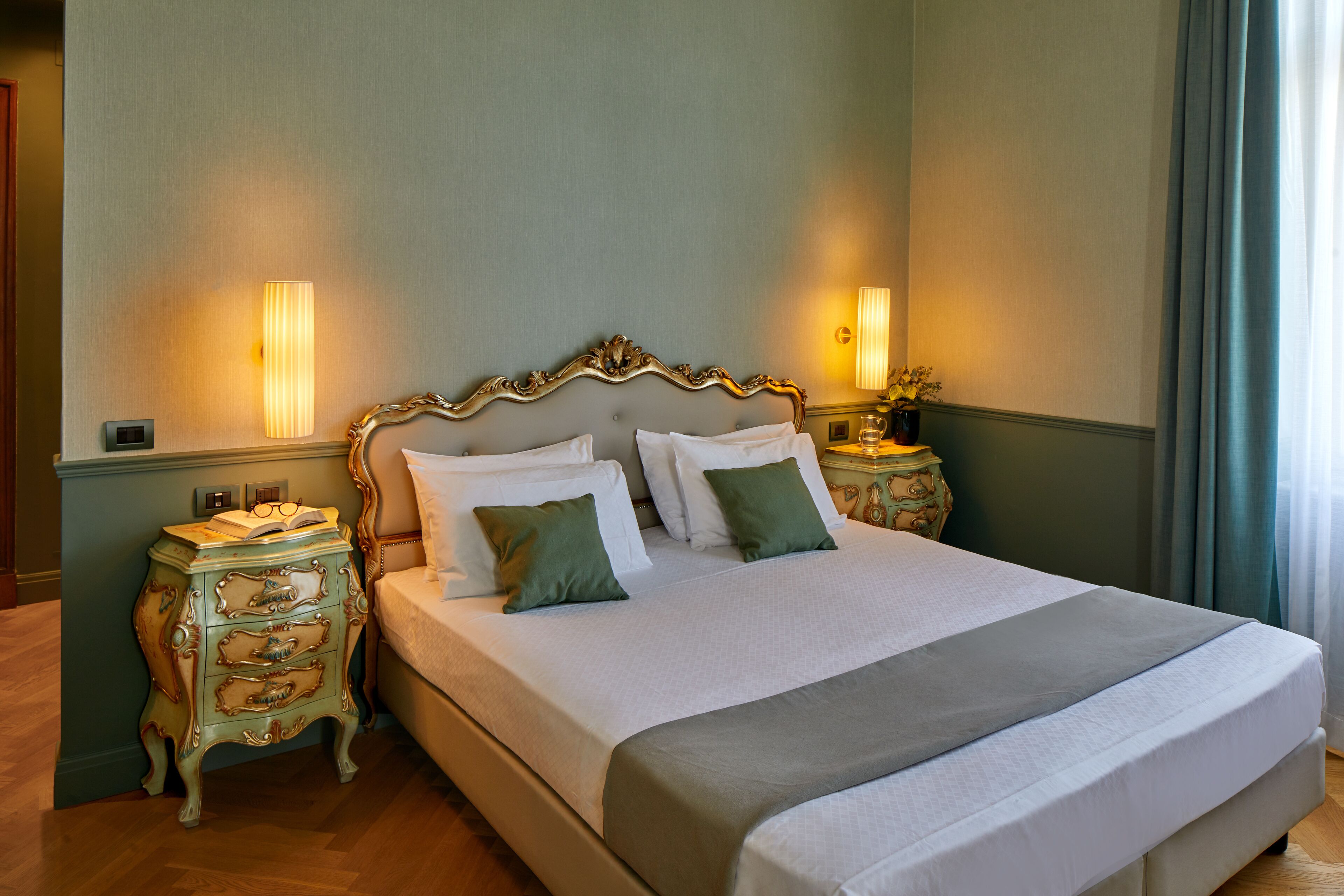 A neatly made bed with a white and gray bedspread, flanked by two nightstands with lamps and a decorative chest of drawers.