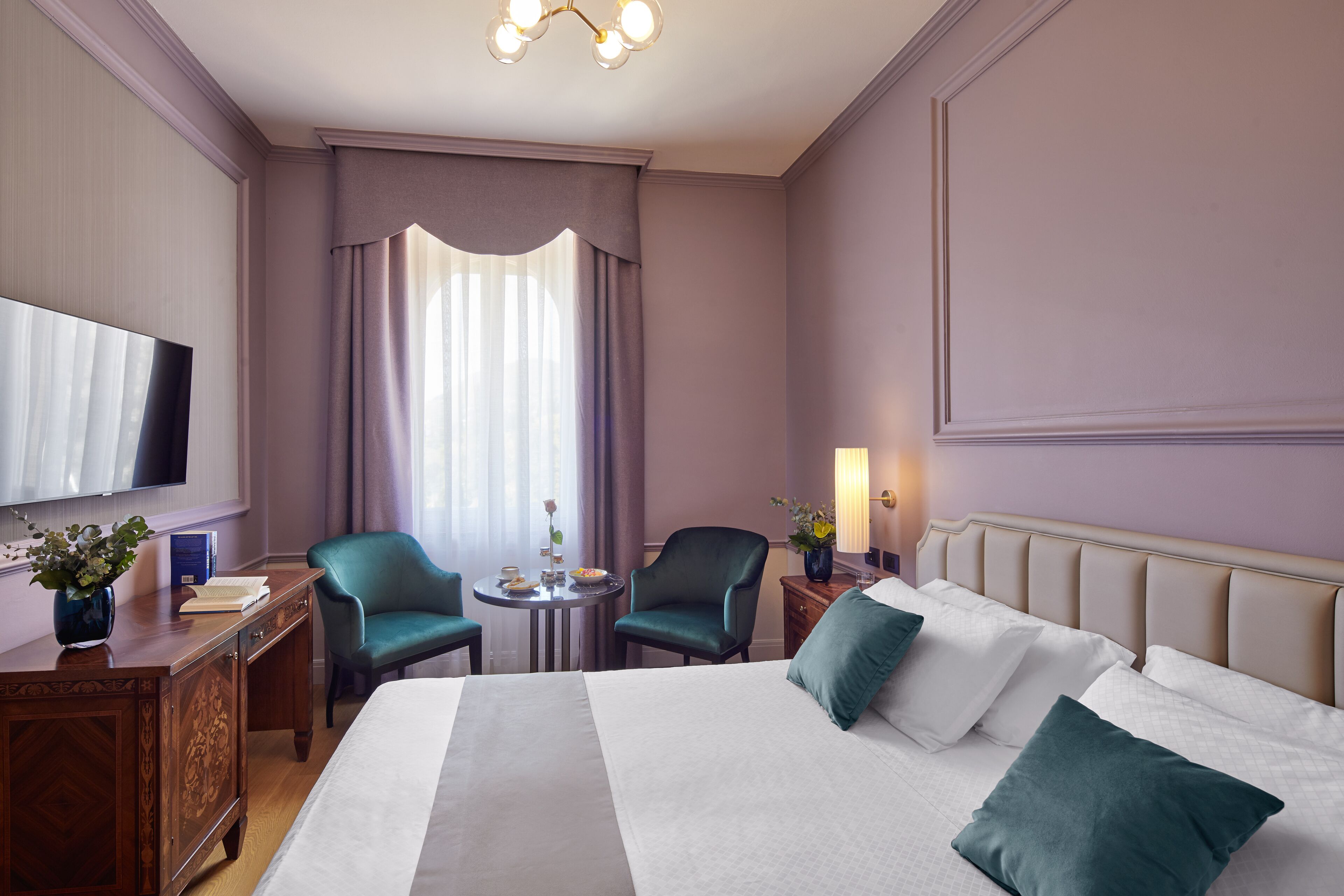 Une chambre d’hôtel avec un grand lit, deux chaises, une petite table avec un vase, une télévision et une lampe.