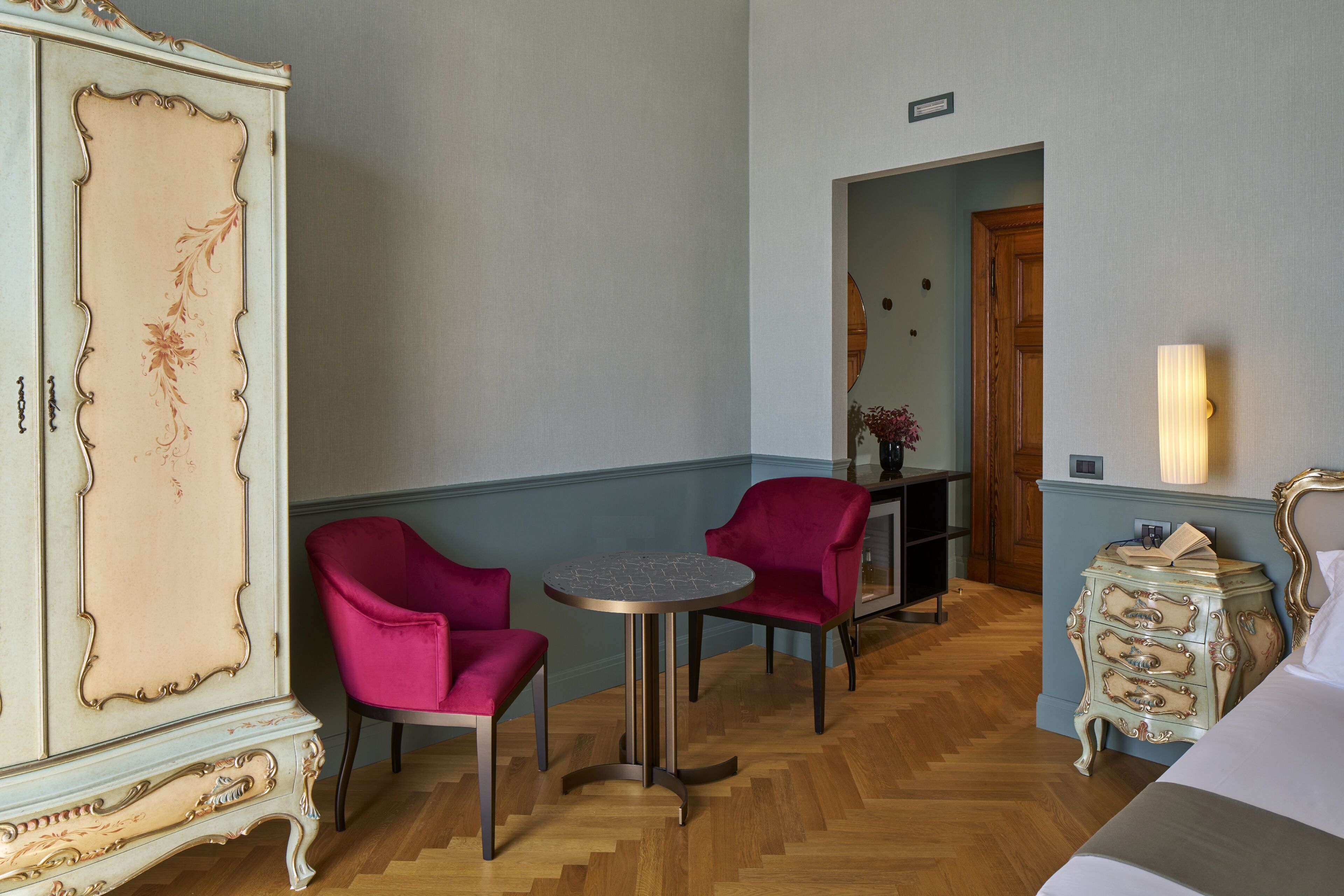 A room with a large ornate cabinet, two red chairs, a small round table, and a dresser with drawers.