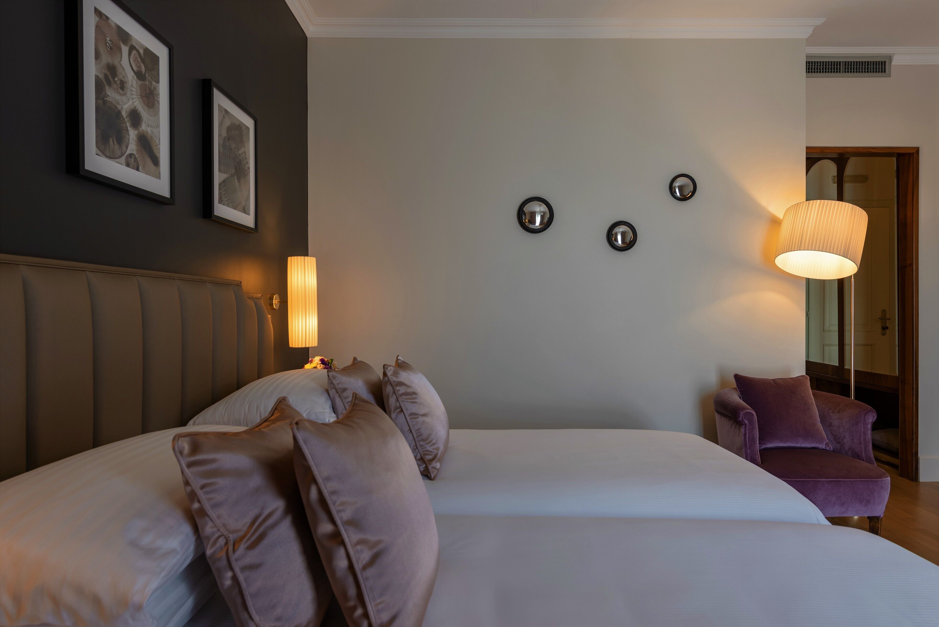 Une chambre d’hôtel avec un grand lit, une tête de lit, un fauteuil violet et des appliques murales.