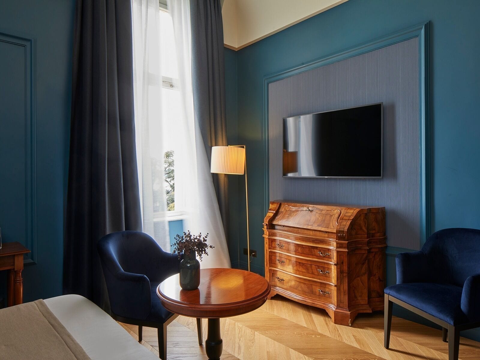 Hotelkamer met een grote flatscreen televisie, twee blauwe fauteuils, een houten dressoir, een ronde houten tafel met een vaas, en een raam met dunne gordijnen.