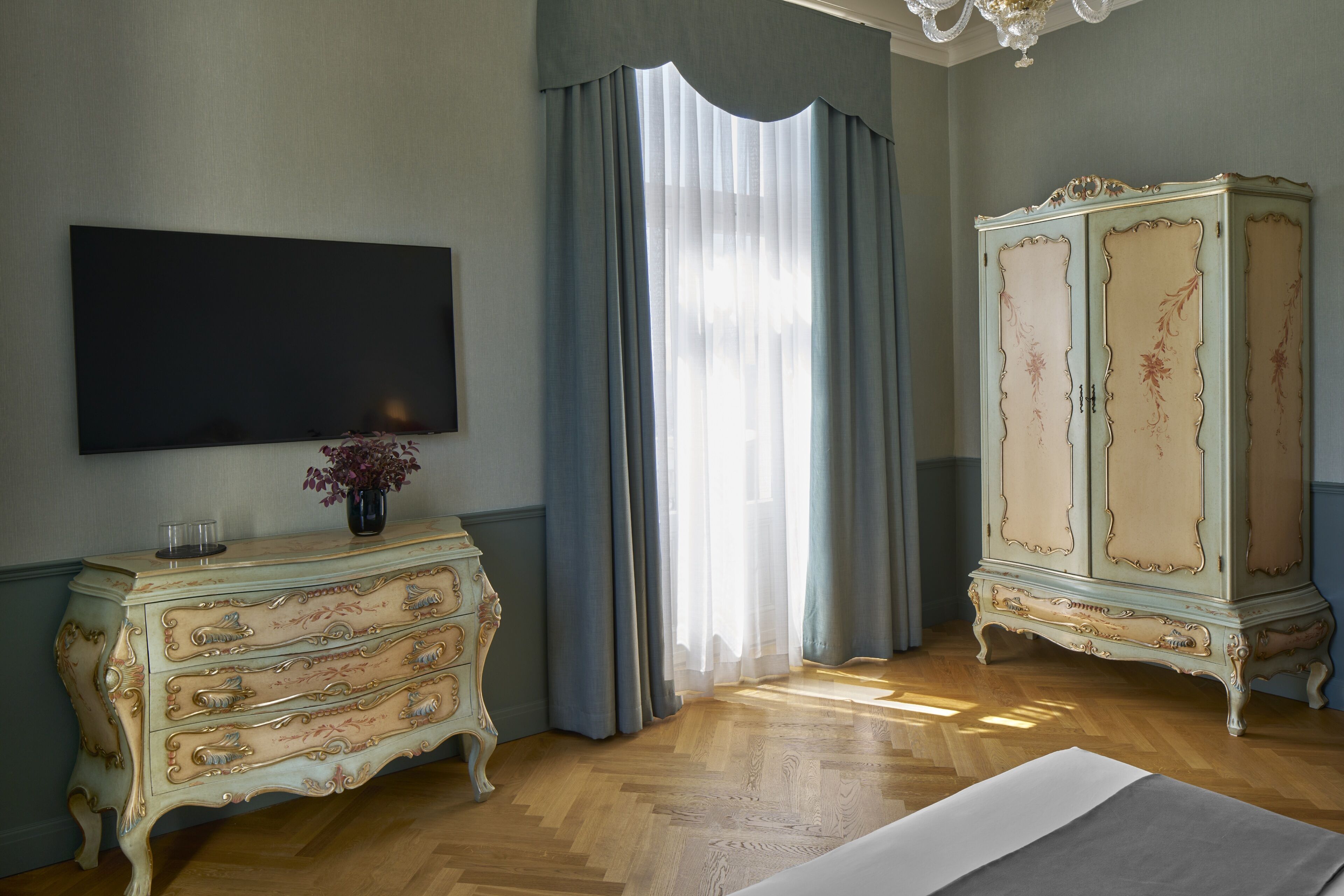 Una camera da letto con pavimento in legno, un comò con motivi floreali, una televisione montata a parete e un'ampia finestra con tende.