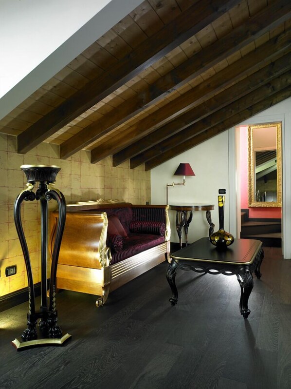 A room with a high ceiling, exposed wooden beams, a red upholstered sofa, a black table with a vase, and a lamp.