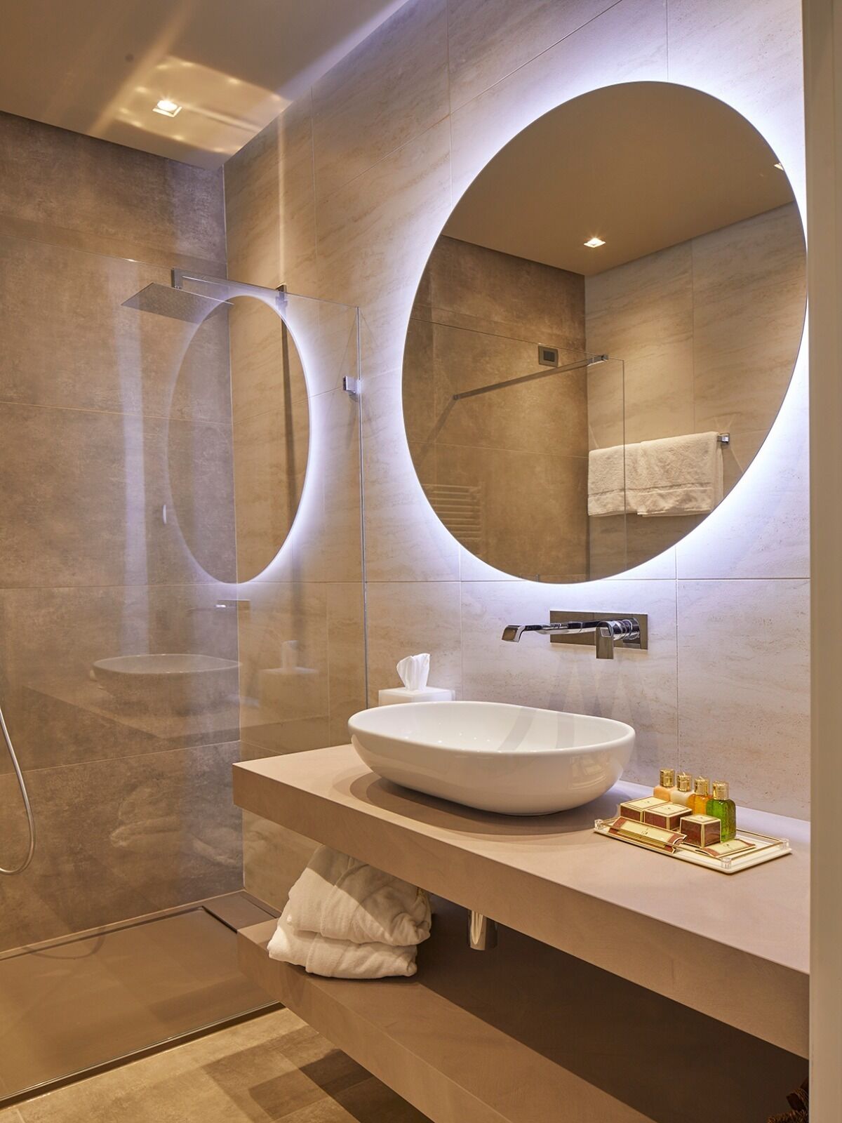 Un baño moderno con un espejo ovalado grande, un lavabo redondo y una ducha con cerramiento de vidrio.