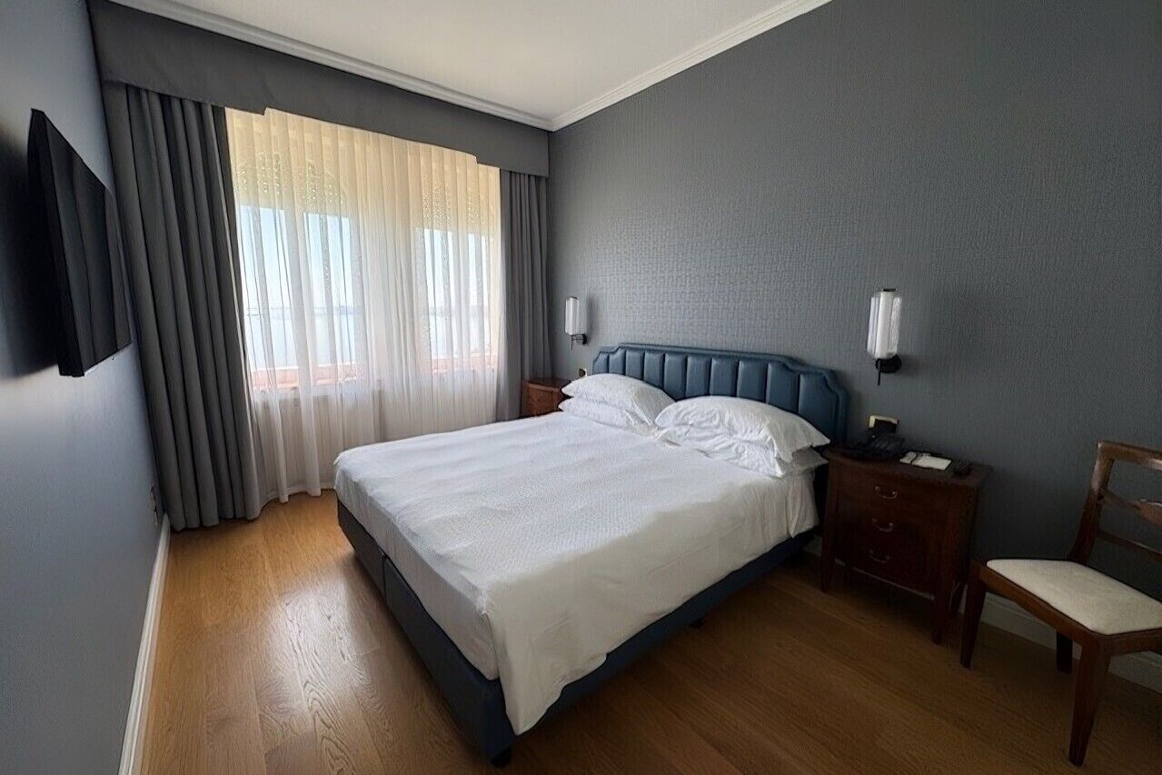 Une chambre à coucher comprenant un lit, une table de chevet, une chaise, un téléviseur fixé au mur et une fenêtre avec des rideaux.