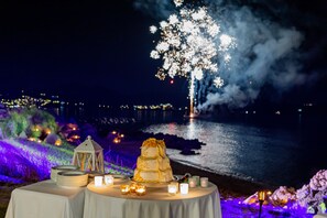 Una mesa con un pastel de bodas de varios niveles, velas y un farol, con fuegos artificiales y una vista al mar de fondo.