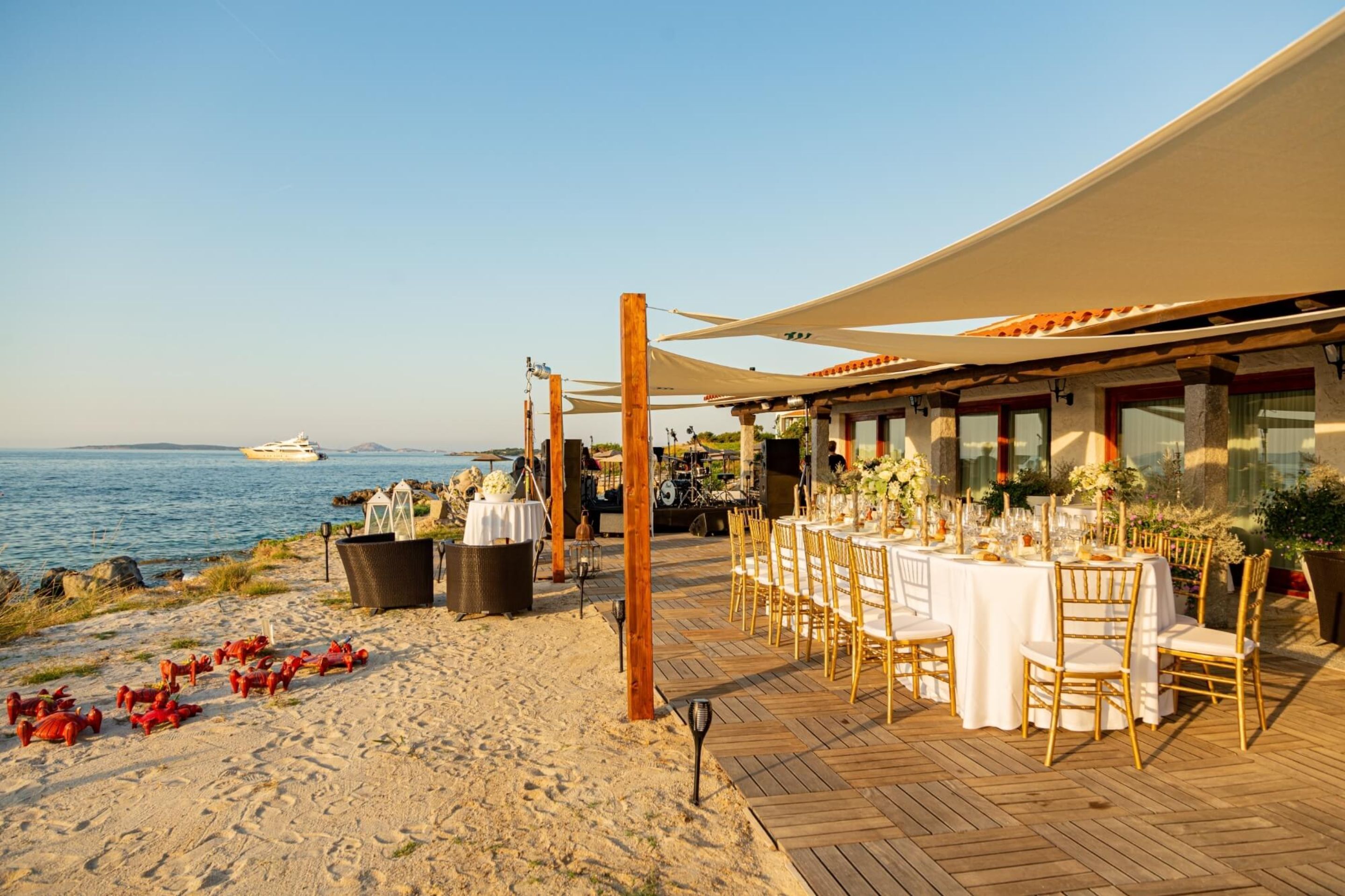 A beachside restaurant with outdoor seating, a view of the sea, and a boat in the background.