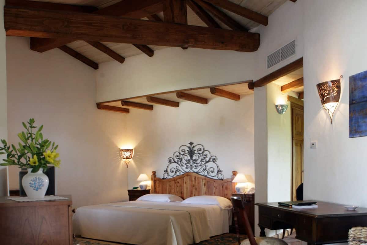 Une chambre avec un plafond en bois, un lit recouvert de draps blancs, une table de chevet en bois, une chaise, un bureau et un miroir fixé au mur.