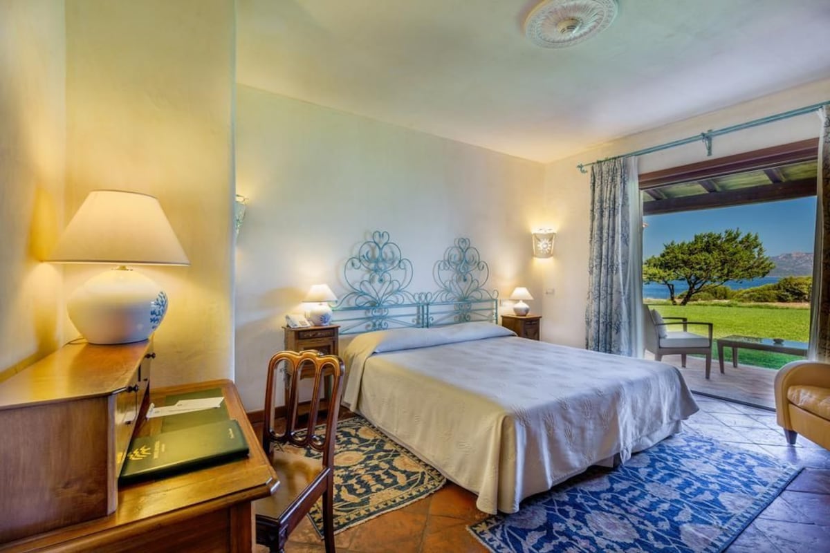 A hotel room with a bed, bedside lamps, a desk with a lamp, a chair, and a view of the outdoors.