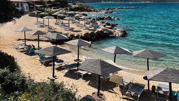 A beach with sun loungers and umbrellas, a rocky shoreline, and mountains in the background. - Hotel Sporting (Olbia)