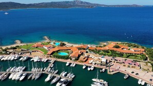An aerial view of a coastal marina with multiple boats docked, a swimming pool, and a complex of buildings with red roofs. - Hotel Sporting (Olbia)