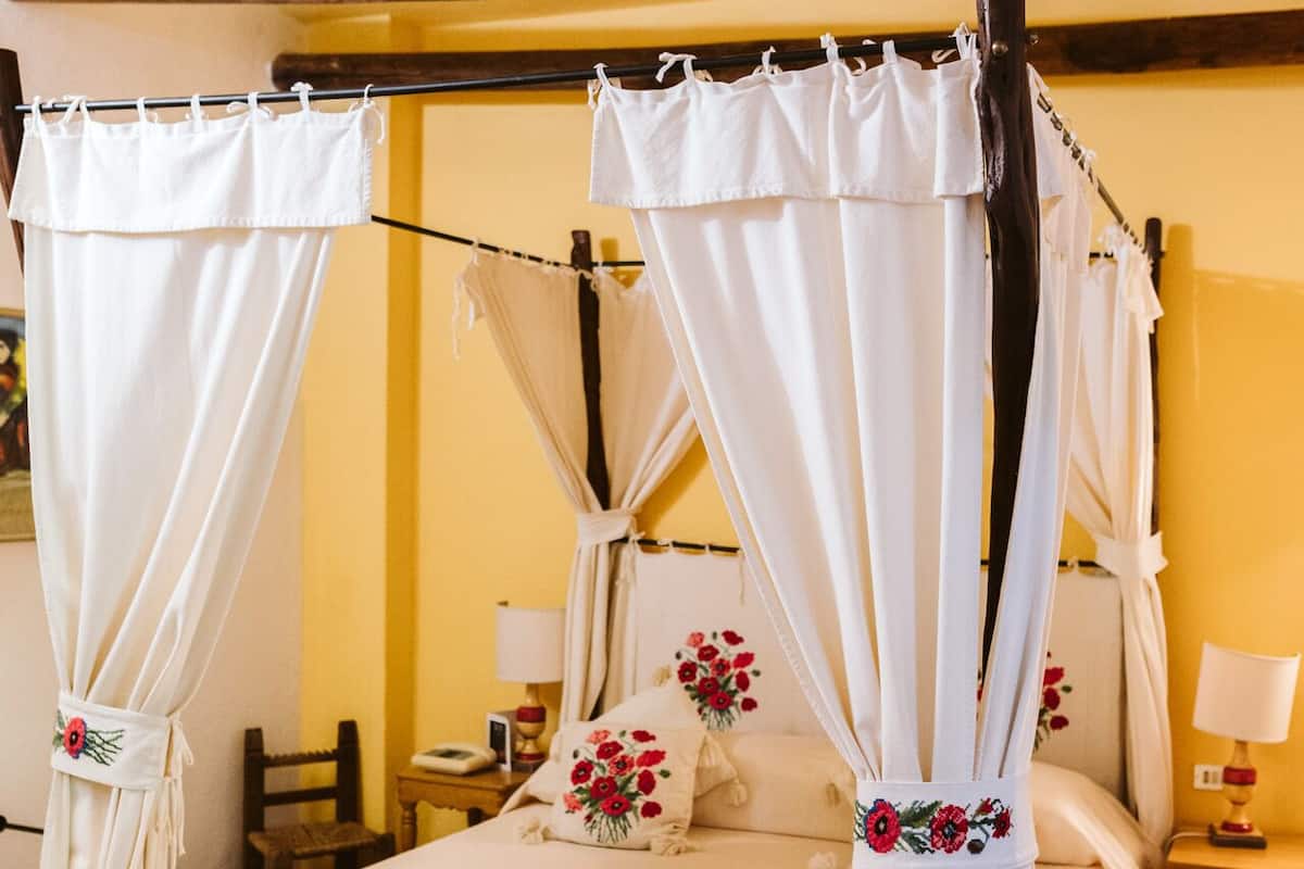 Un lit à baldaquin avec du linge de lit blanc et des coussins à motifs floraux, un plafond en bois et un mur jaune.