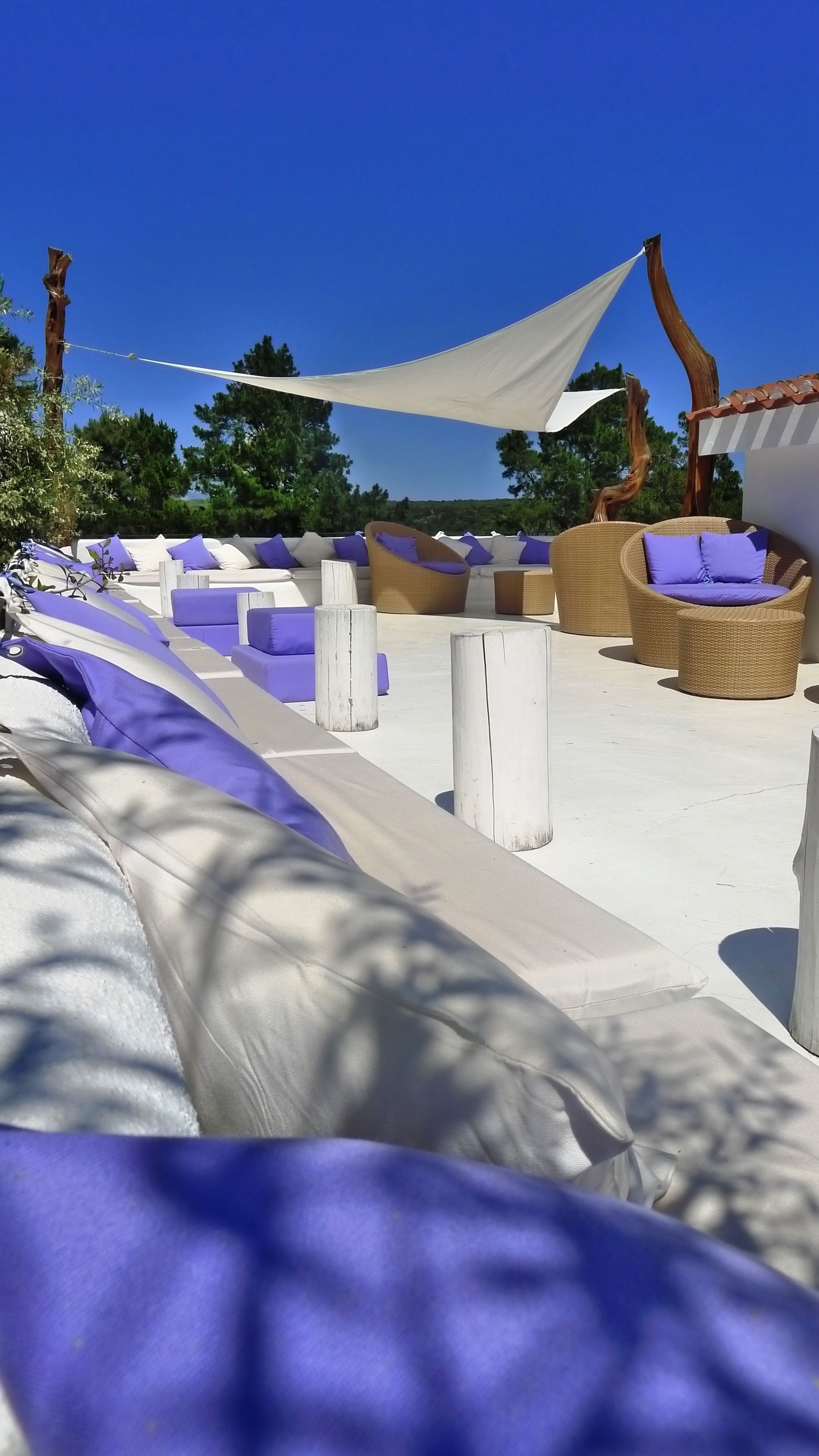 A sunlit outdoor seating area with white and purple cushions, wooden posts, and a white canopy.