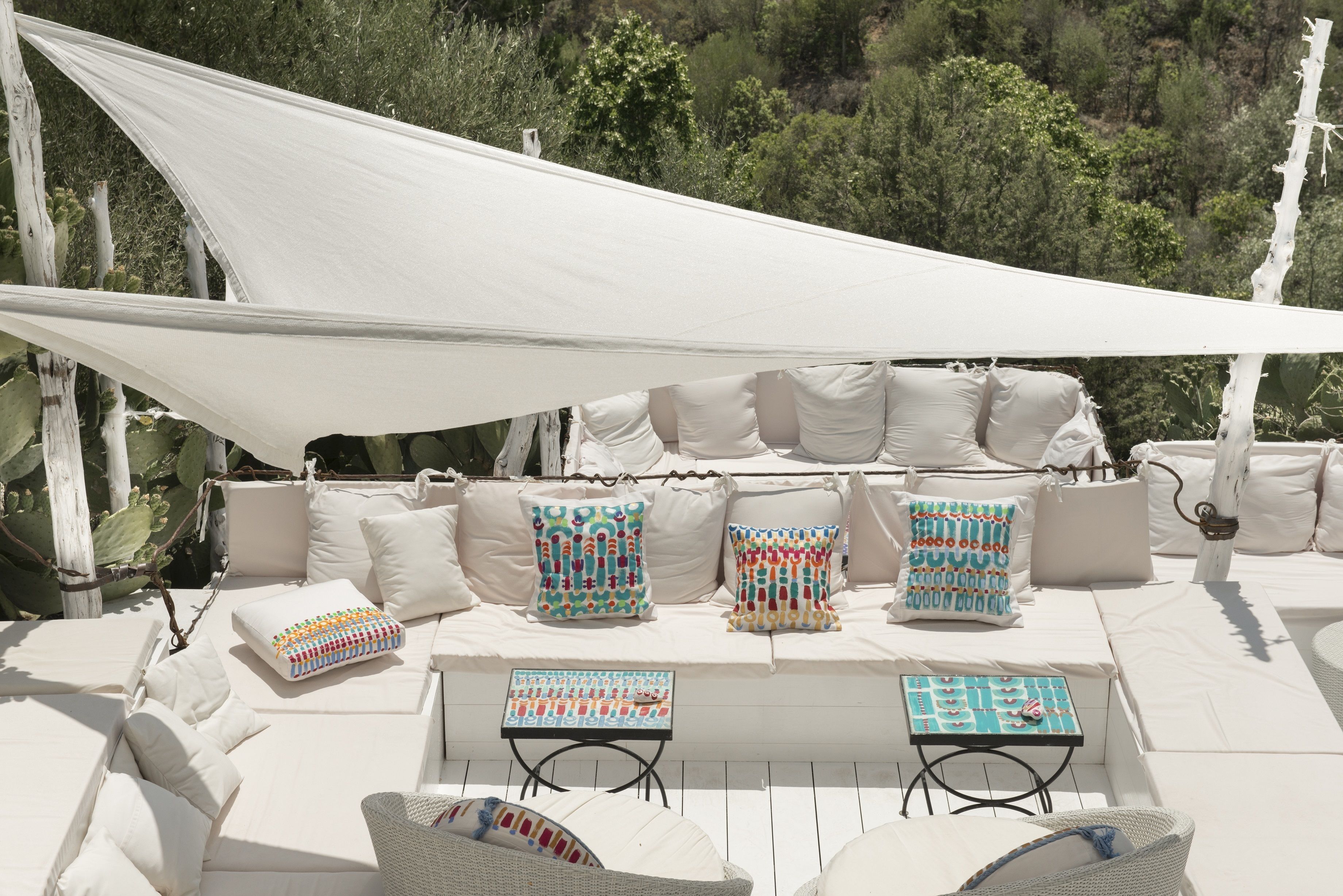 A sunlit outdoor seating area with white cushions, colorful pillows, and two small tables.