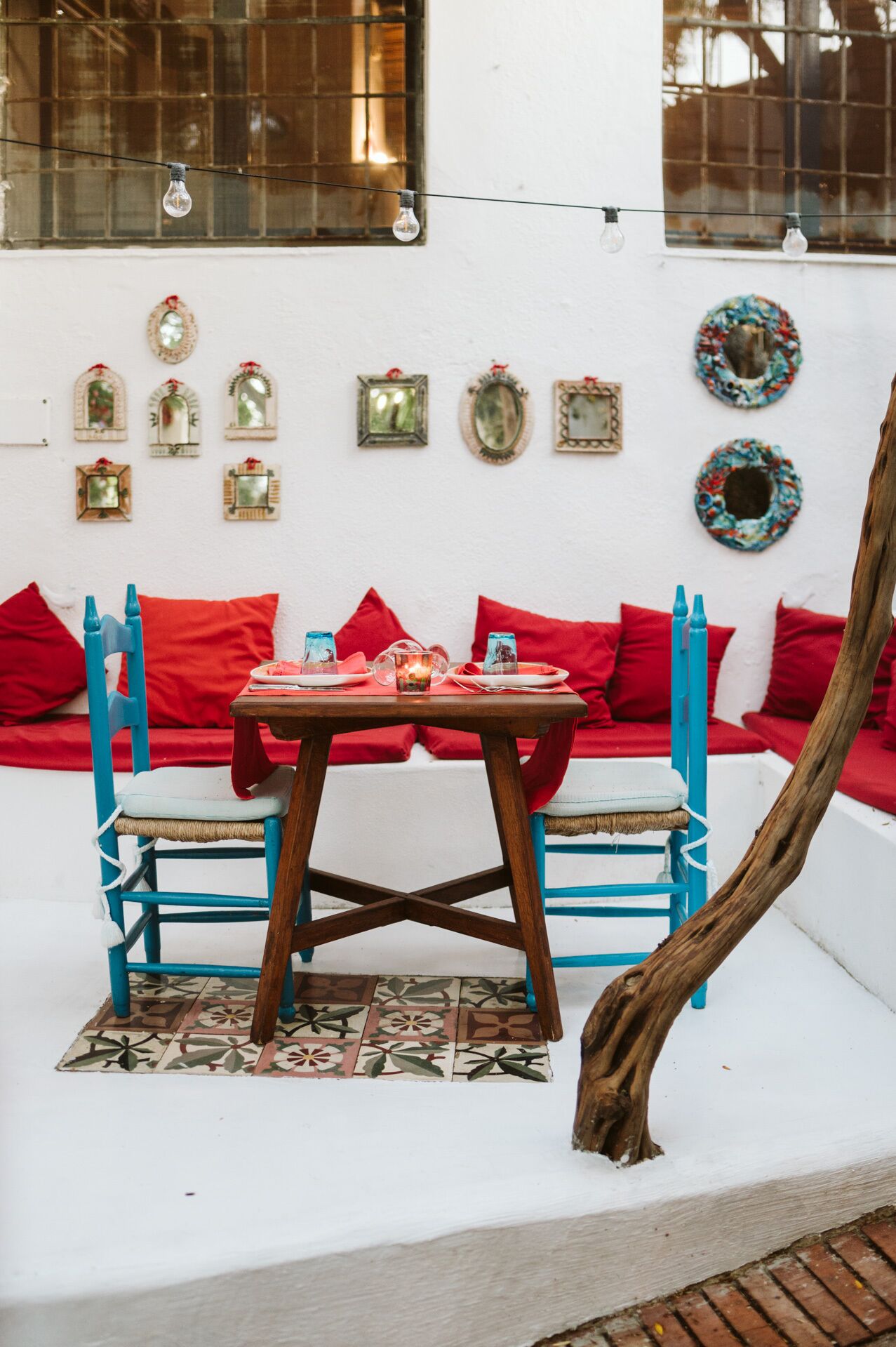 Un rincón al aire libre acogedor, con una mesa preparada para dos, rodeado de cojines rojos intensos y sillas azules.