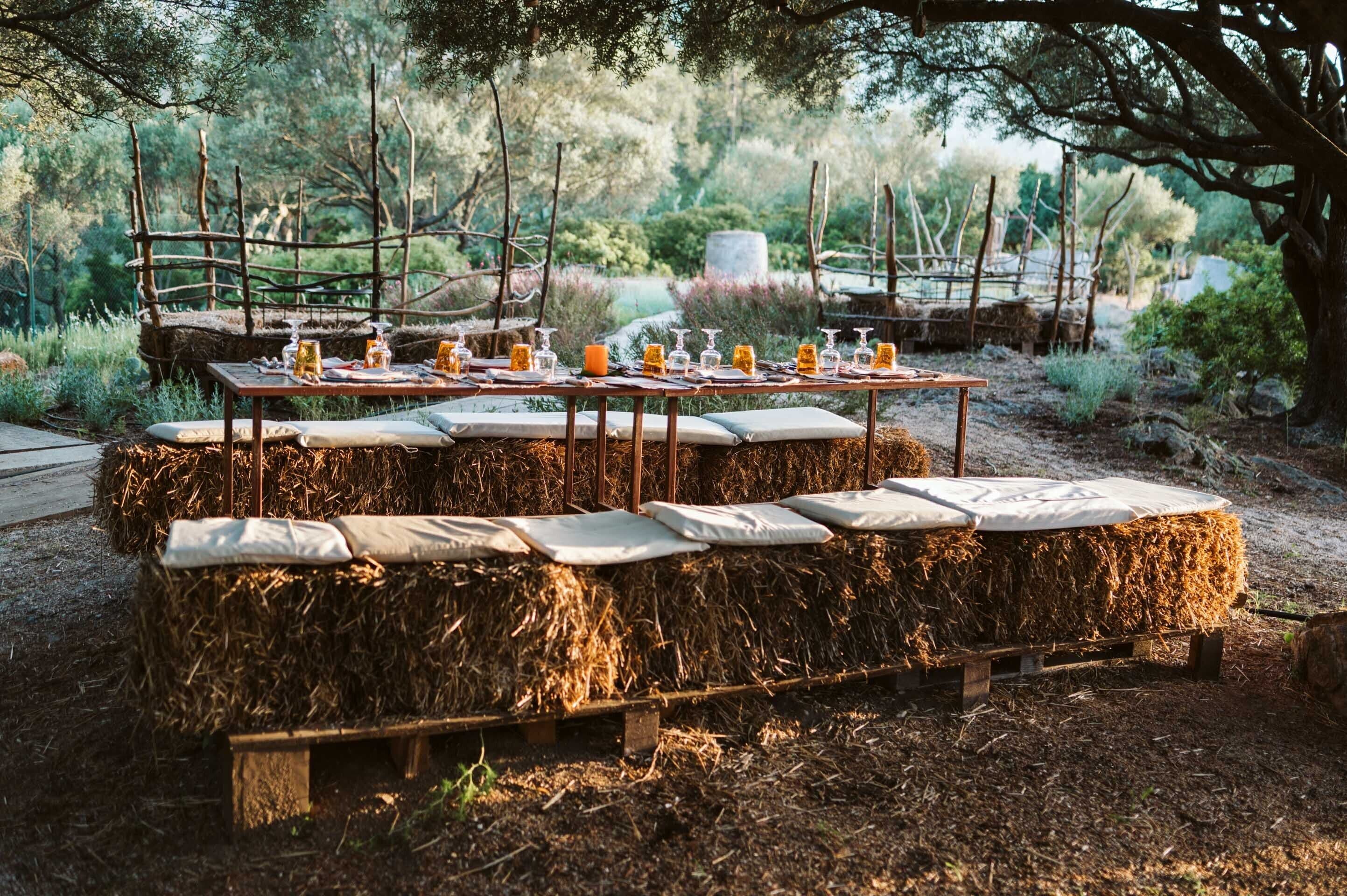 A rustic outdoor dining setup with hay bales and wooden tables, set in a natural, wooded area.