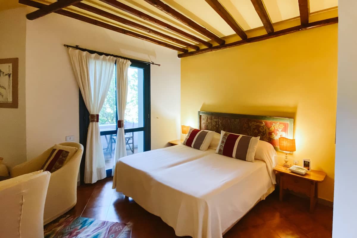 Une chambre d’hôtel avec un grand lit, une chaise, une petite table avec une lampe et une fenêtre avec des rideaux.