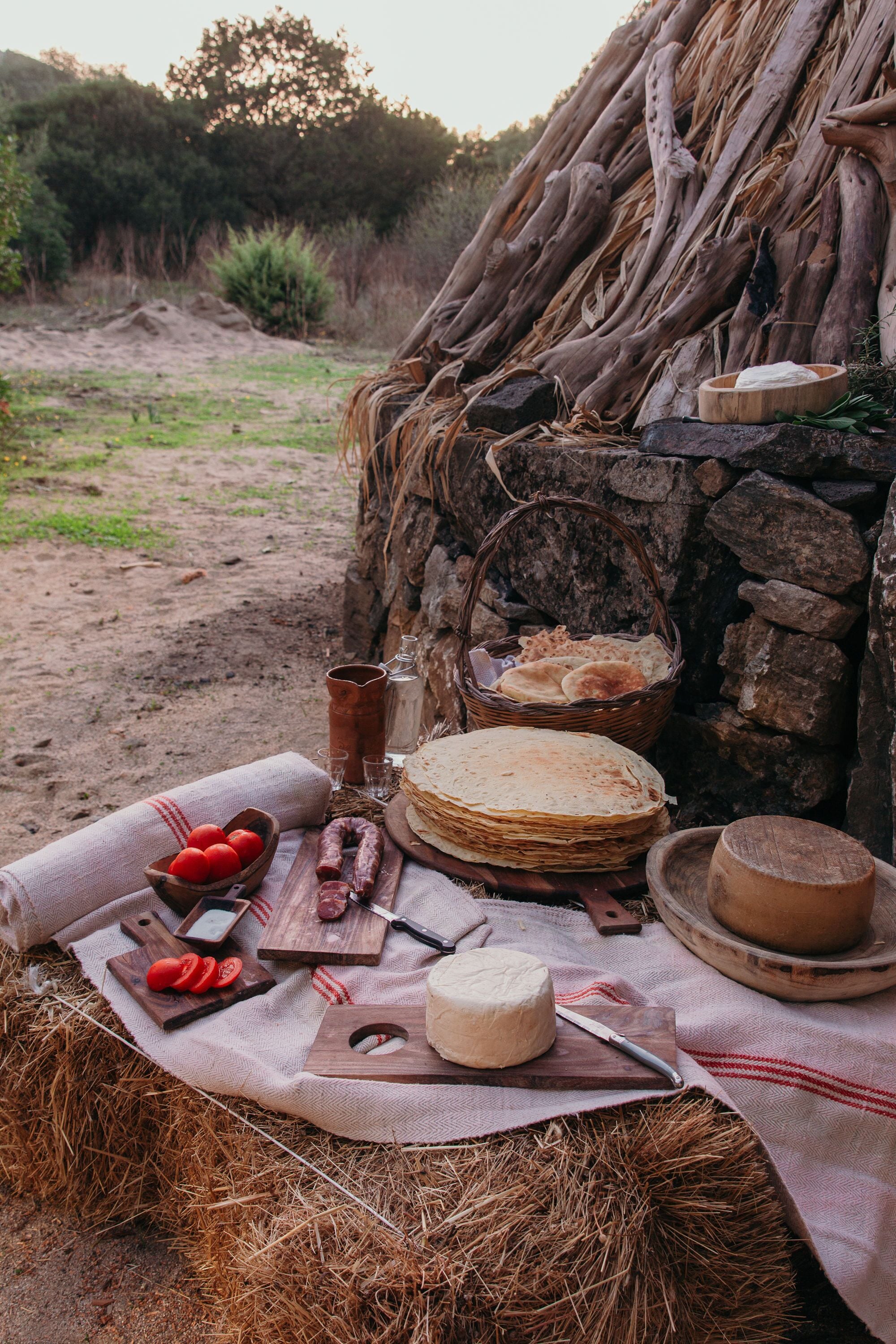 A rustic outdoor dining setup with a woven basket, wooden cutting board, cheese, and bread on a straw-covered table.