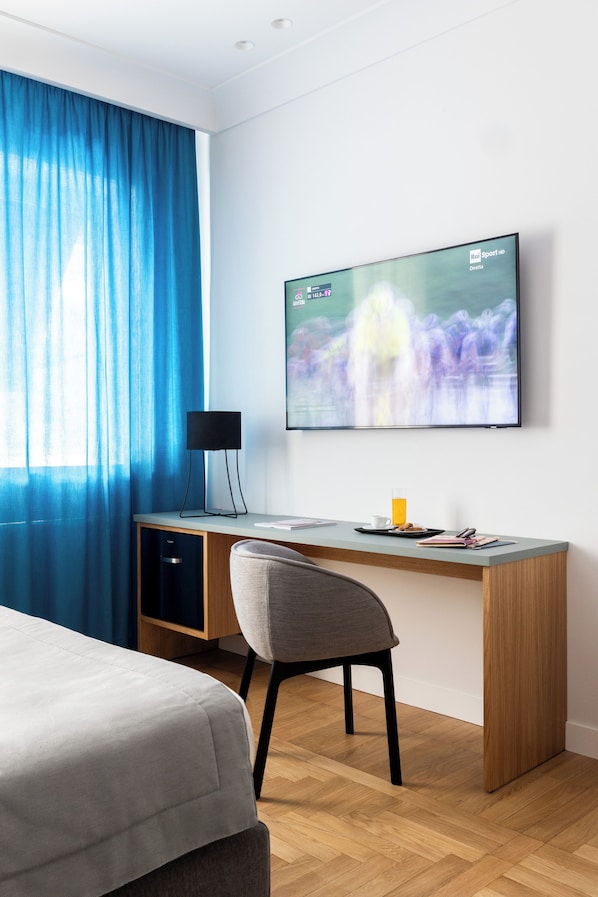 A modern hotel room with a bed, a desk, a chair, a TV, and a window with blue curtains.