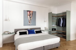Una camera da letto moderna con un letto grande, un comodino con libri e una tazza, un quadro appeso al muro, un armadio con mensole e un appendiabiti.