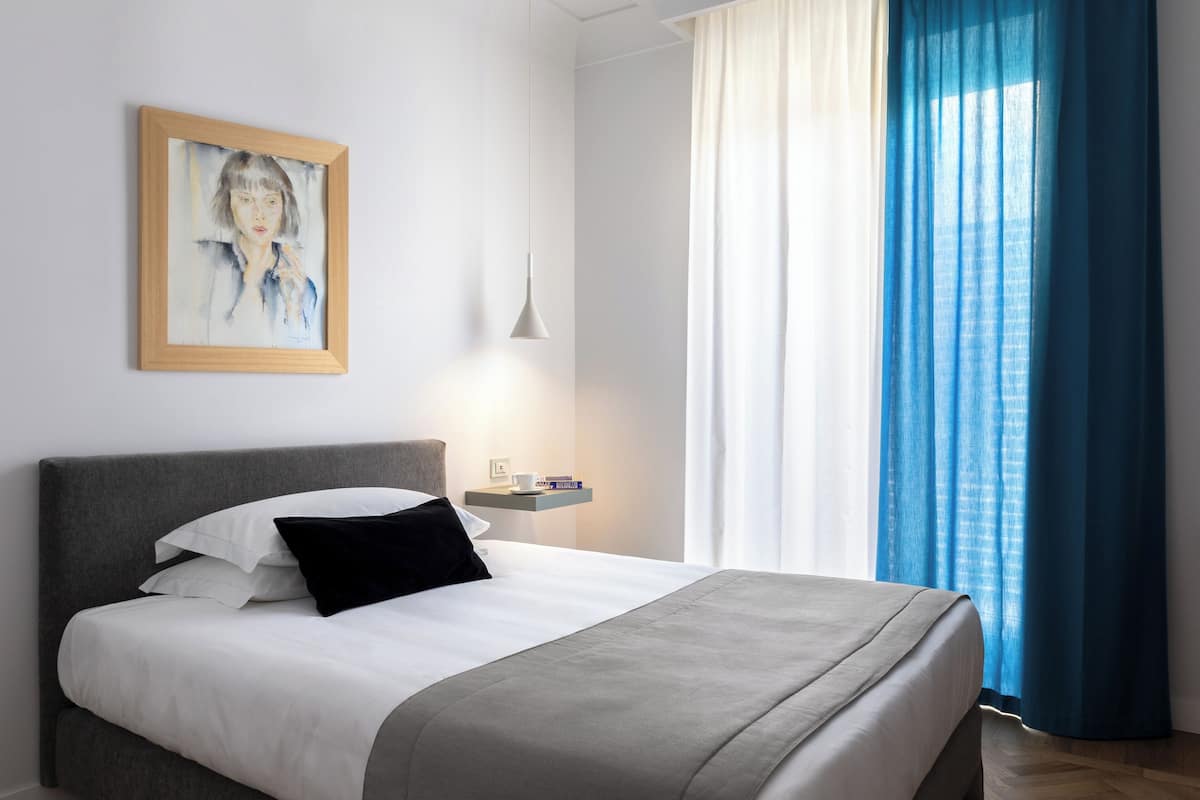 A modern hotel room with a neatly made bed, a bedside table with a lamp, a painting on the wall, and a view through blue curtains.
