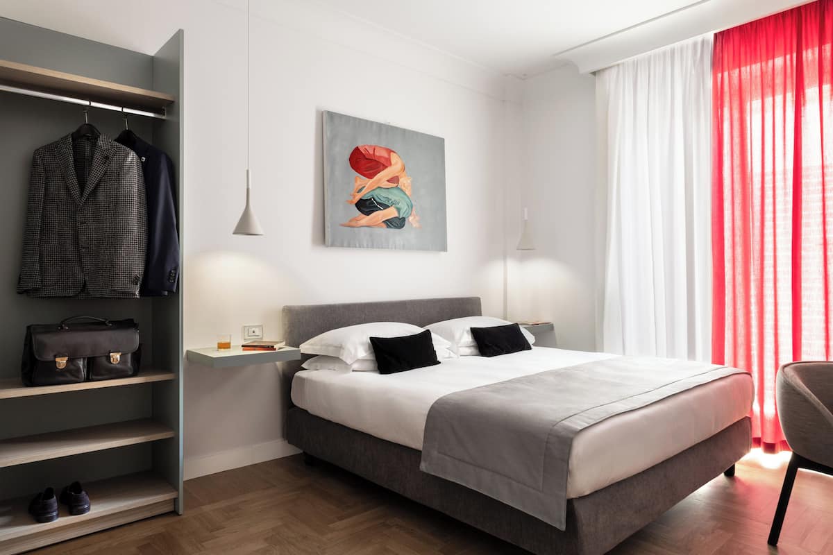 A modern bedroom with a grey bed, white pillows, a grey headboard, a grey wardrobe, a grey bench, and a red curtain.