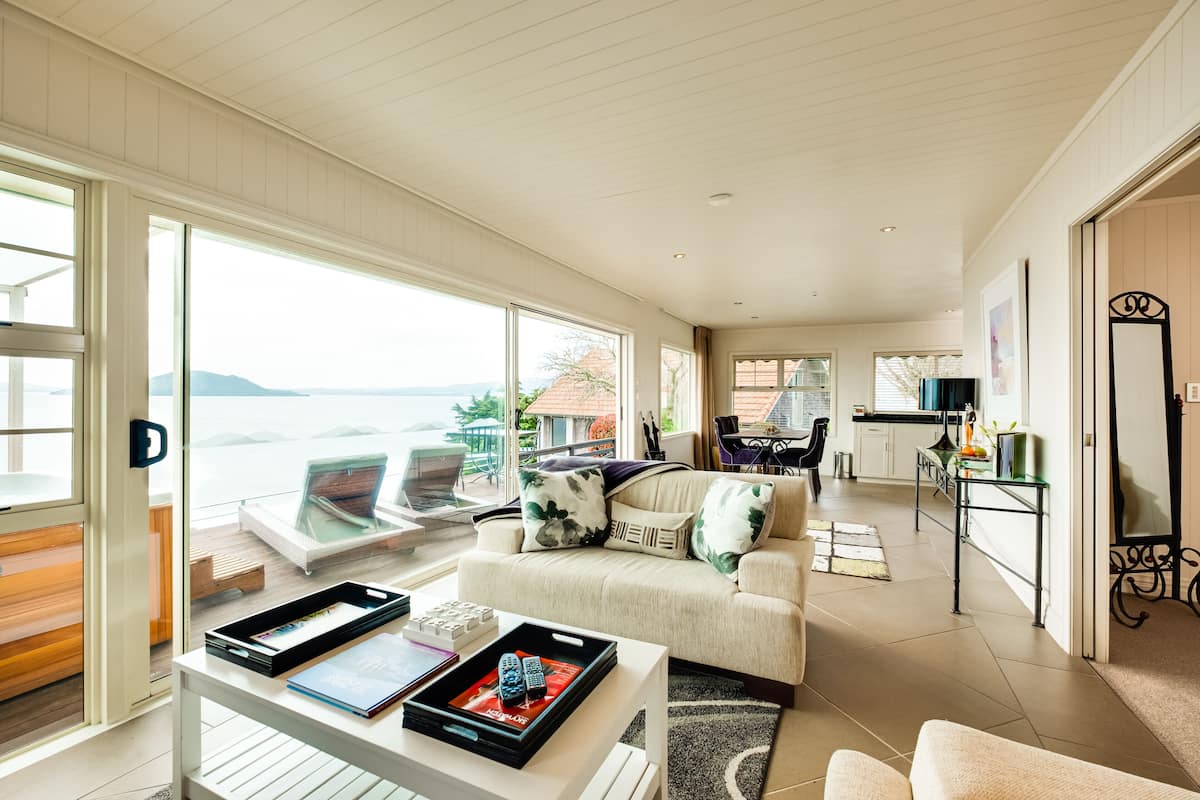 Un amplio salón con un gran ventanal con vistas panorámicas, un rincón de descanso con sofá y sillones, y una cocina al fondo.