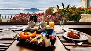 A breakfast table with a variety of fruits, a croissant, and a glass of orange juice, set against a scenic view of a lake and mountains. - On The Point - Lake Rotorua (Rotorua)