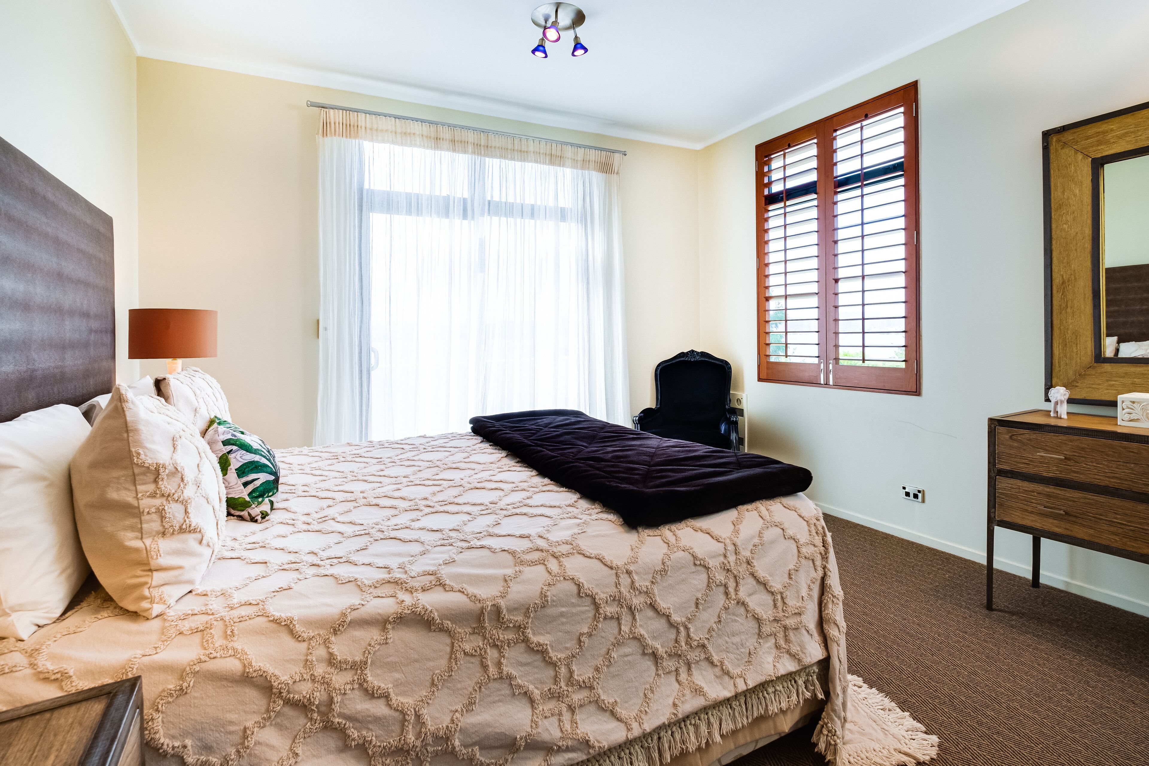 A neatly arranged bedroom with a bed, a bedside lamp, a dresser, and a window with blinds.