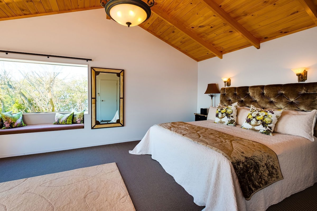 Un dormitorio con una cama grande, un rincón con asiento junto a la ventana, un espejo y una lámpara en el techo.