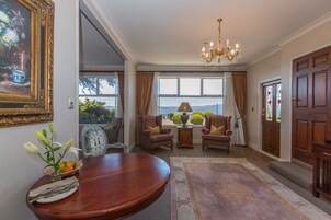 Uma sala de estar formal com uma mesa redonda de madeira, duas poltronas, um candelabro e vista através de amplas janelas.