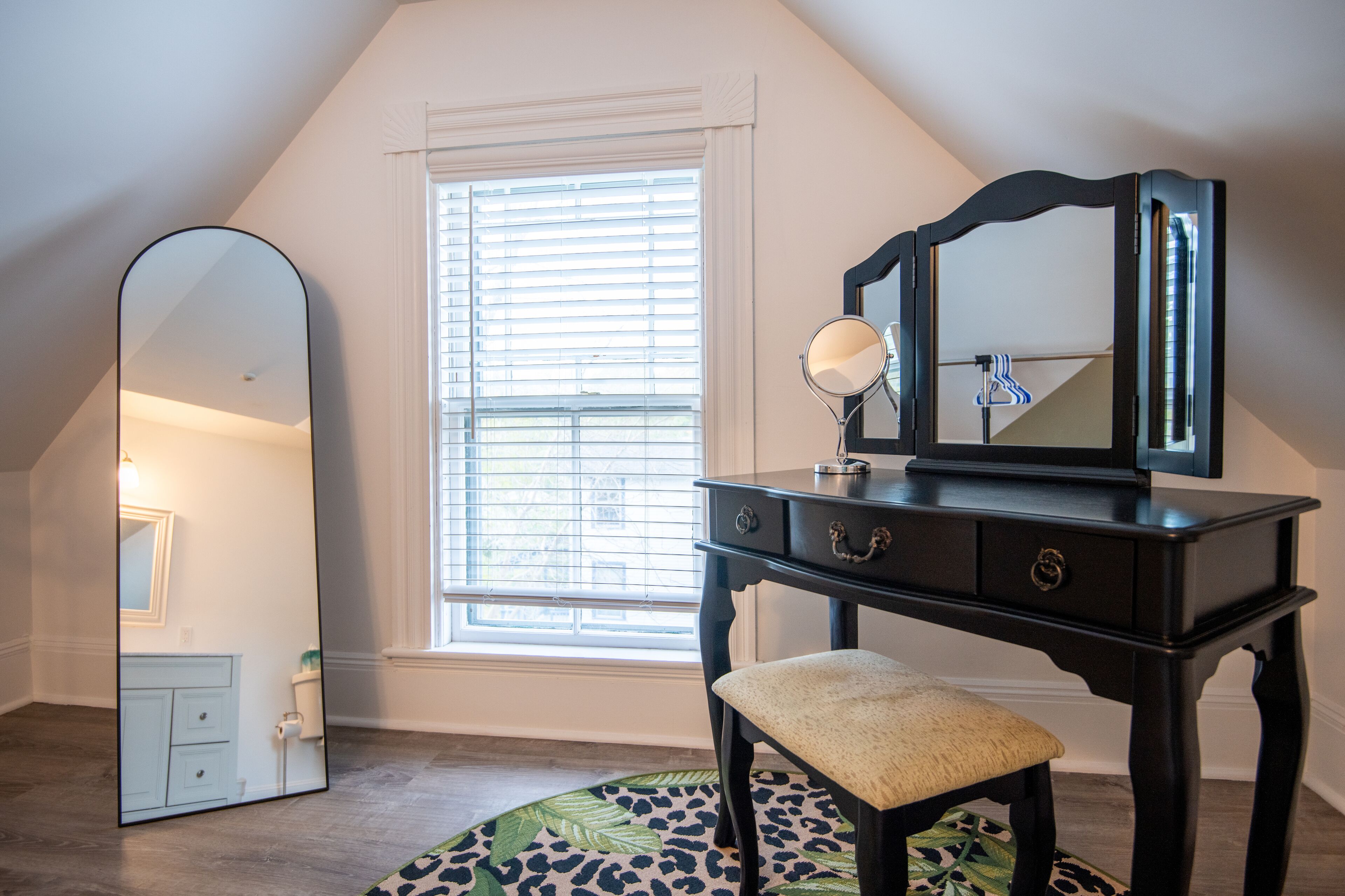 A room with a vanity, a large mirror, a window with blinds, and a leopard print rug.