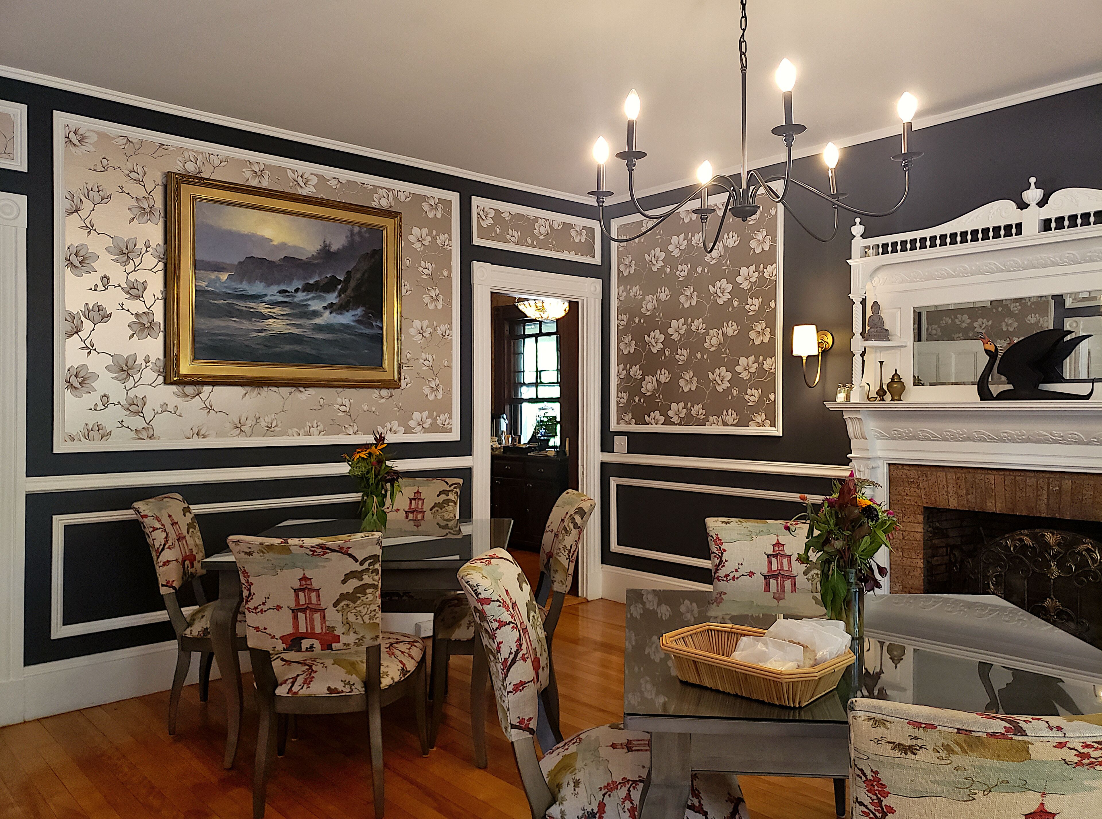 Uma sala de jantar com mesa, cadeiras e um lustre. Há um quadro na parede e uma lareira com um cisne decorativo.