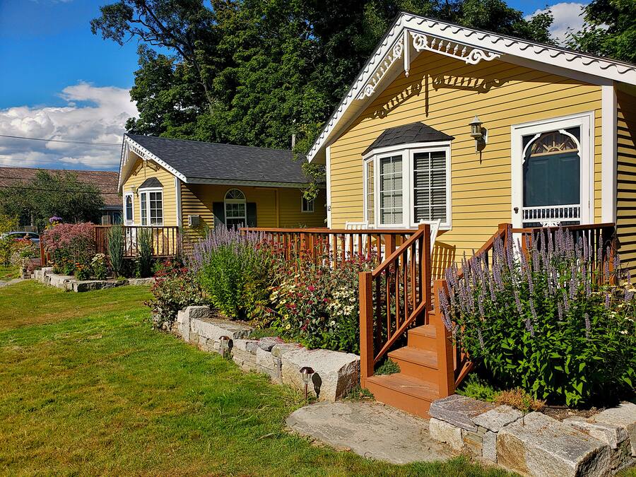 A yellow house with a porch, a stone retaining wall, and a variety of flowers.