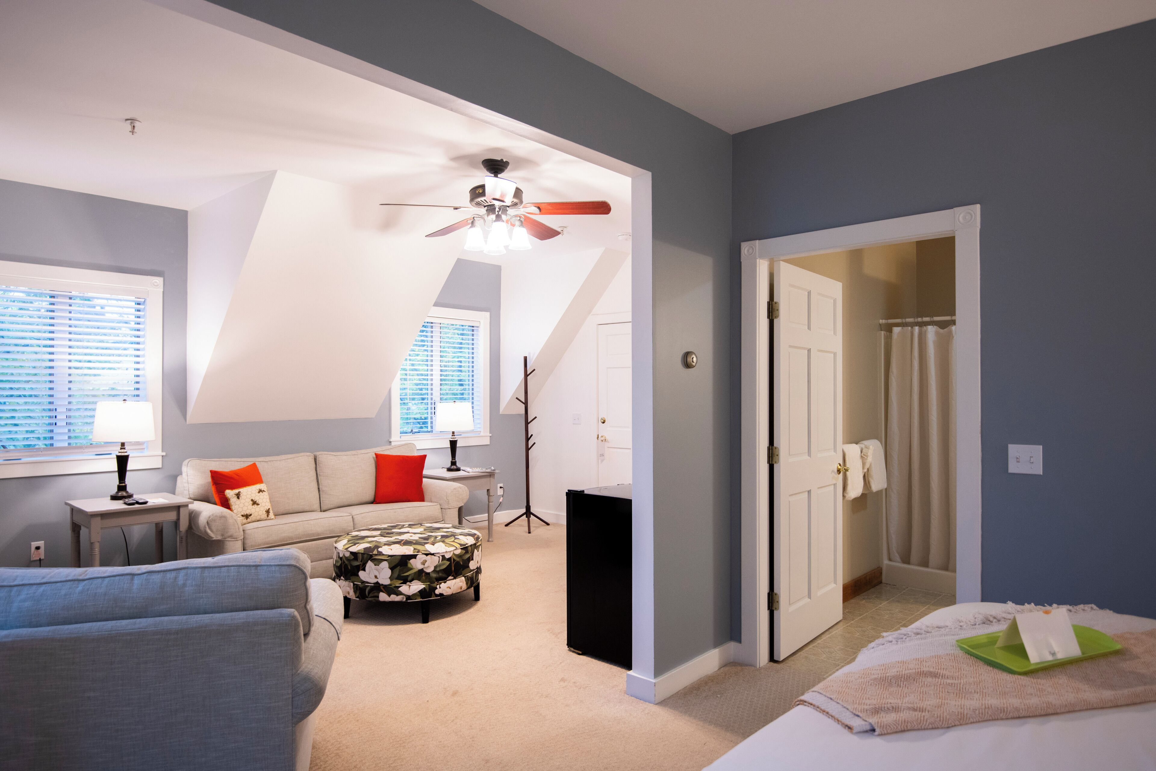 Une chambre à coucher comprenant un lit, une table de chevet, une commode, une fenêtre avec des stores, un ventilateur de plafond et une porte donnant sur une autre pièce.