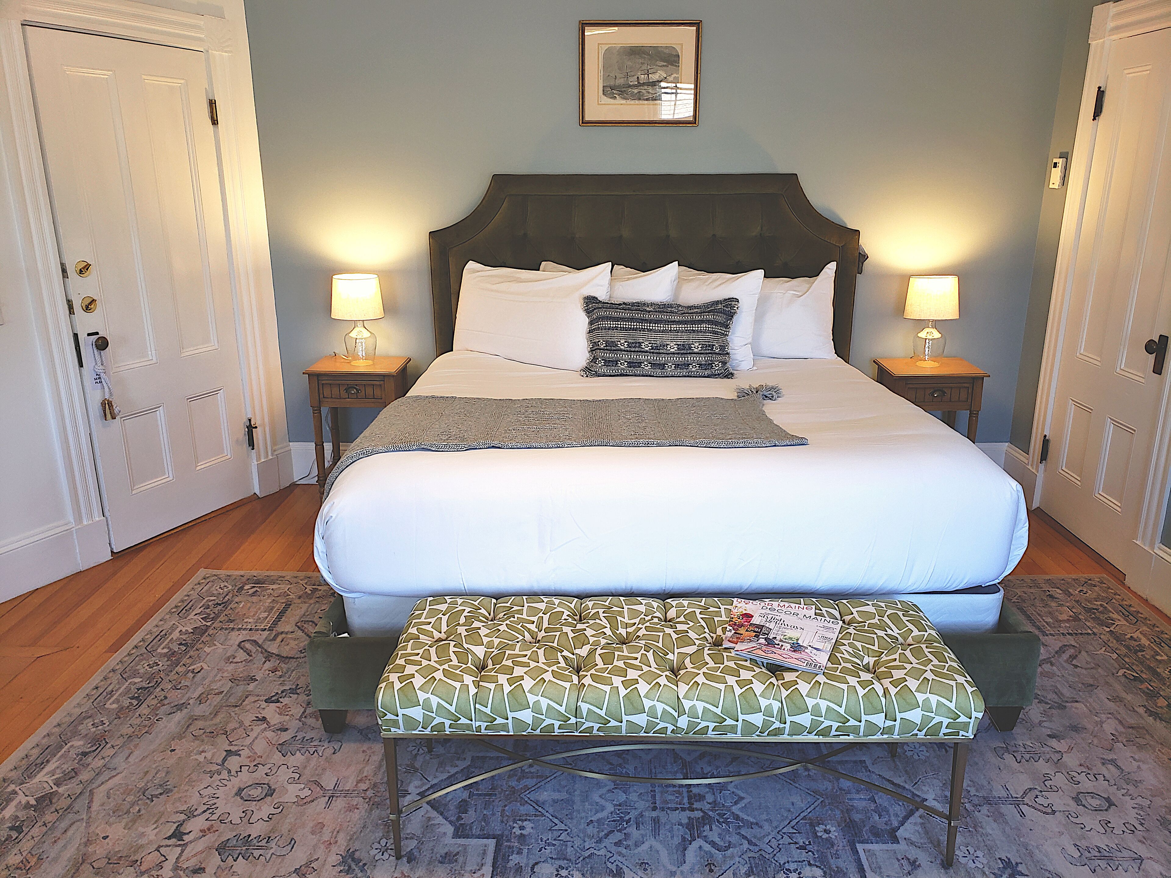 A neatly made bed with a headboard, two bedside lamps, a framed picture above the bed, and a patterned bench in front of the bed.