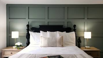Cama bem arrumada com lençóis brancos e uma revista sobre ela. Duas mesas de cabeceira, com abajures e um vaso de flores em uma delas. O quarto tem painel de cabeceira verde e parede verde-clara.