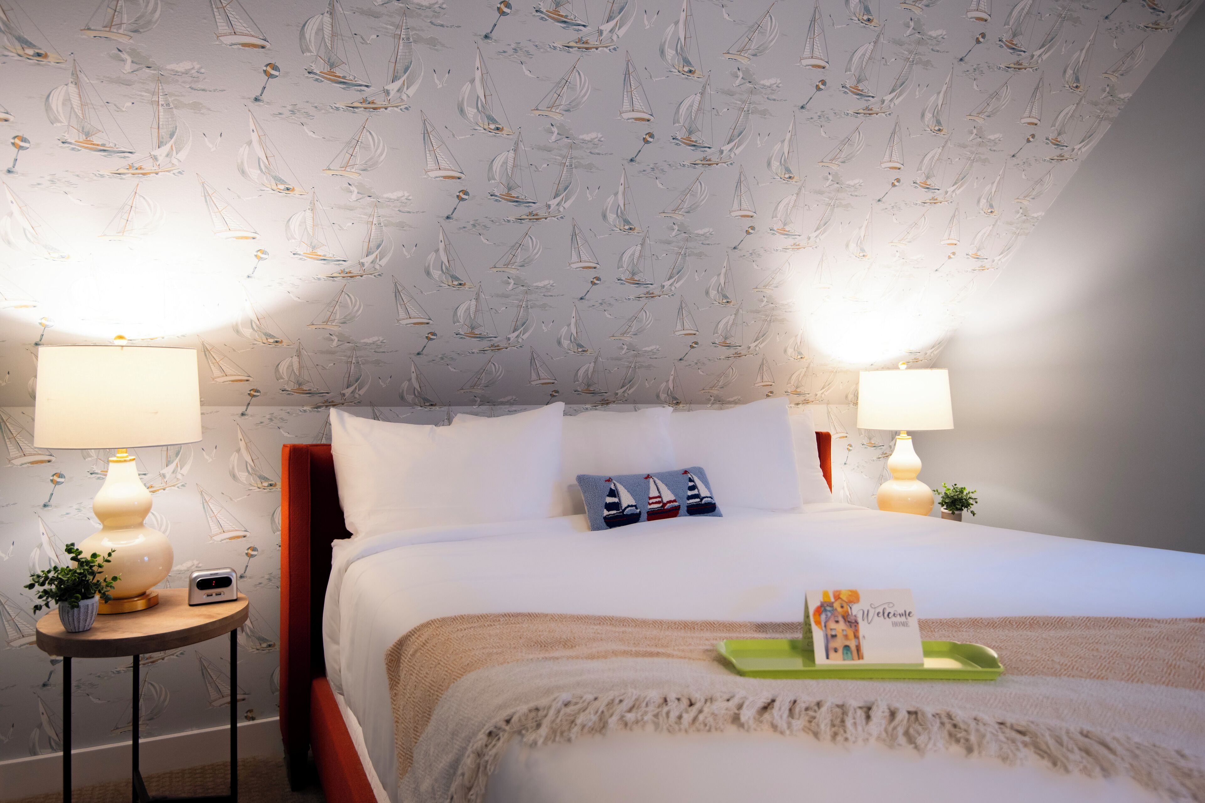 A hotel room with a bed, two bedside lamps, a small table with a lamp, a welcome sign, and a wall with sailboat wallpaper.