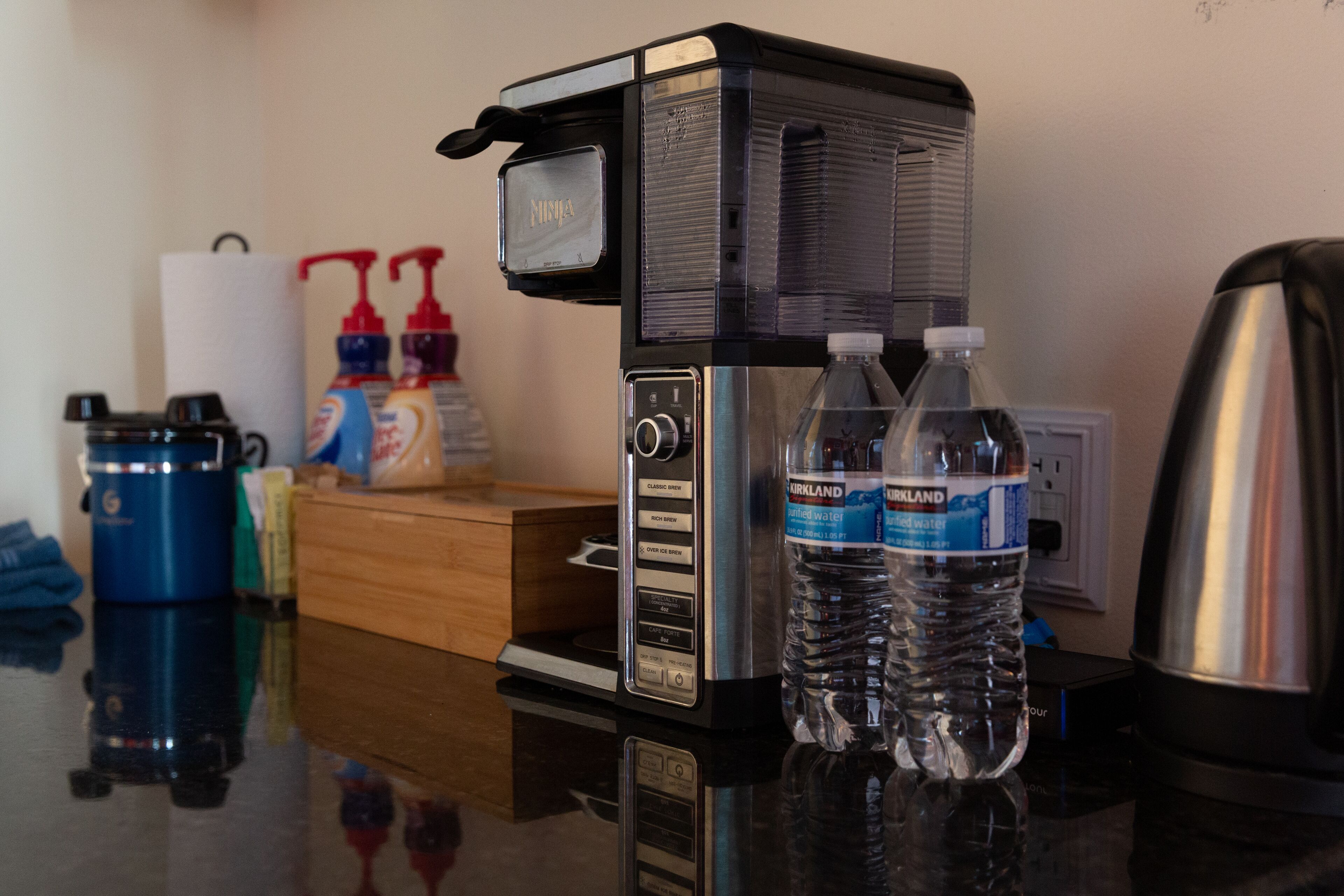A coffee maker, two bottles of Kirkland water, a coffee pot, and a tissue dispenser on a reflective countertop.