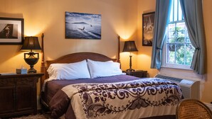 A hotel room with a bed, two bedside tables with lamps, a window with curtains, a wall-mounted air conditioner, and a framed picture above the bed. - Blue60 Marigny Inn (New Orleans)