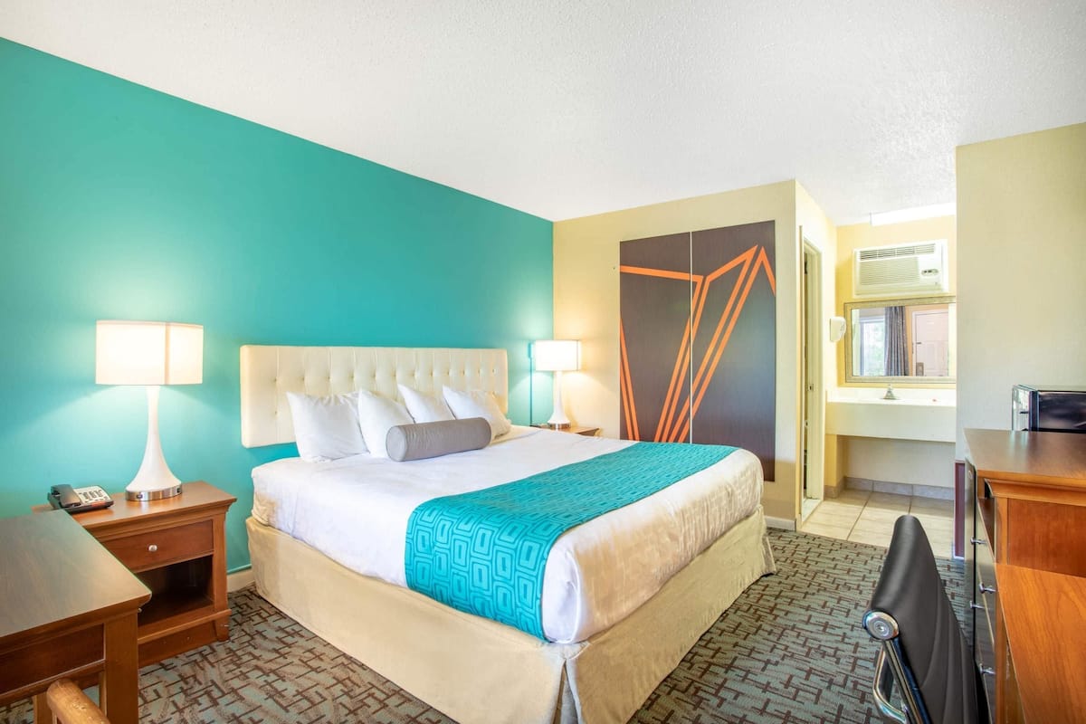 A hotel room with a bed, bedside tables, a desk, a chair, and a bathroom visible through an open door.