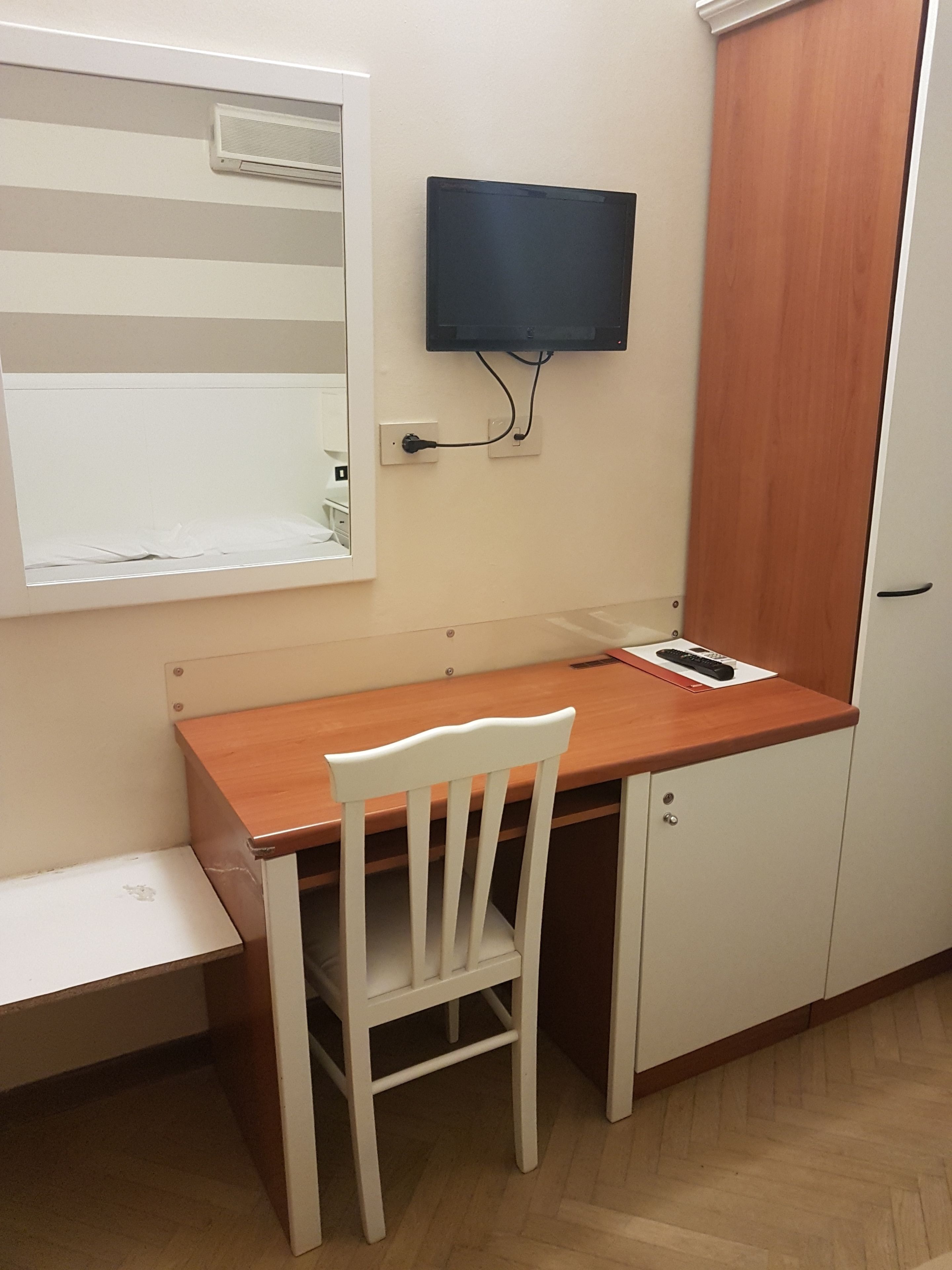 Une petite pièce avec un bureau en bois, une chaise blanche, un téléviseur fixé au mur et une armoire en bois.