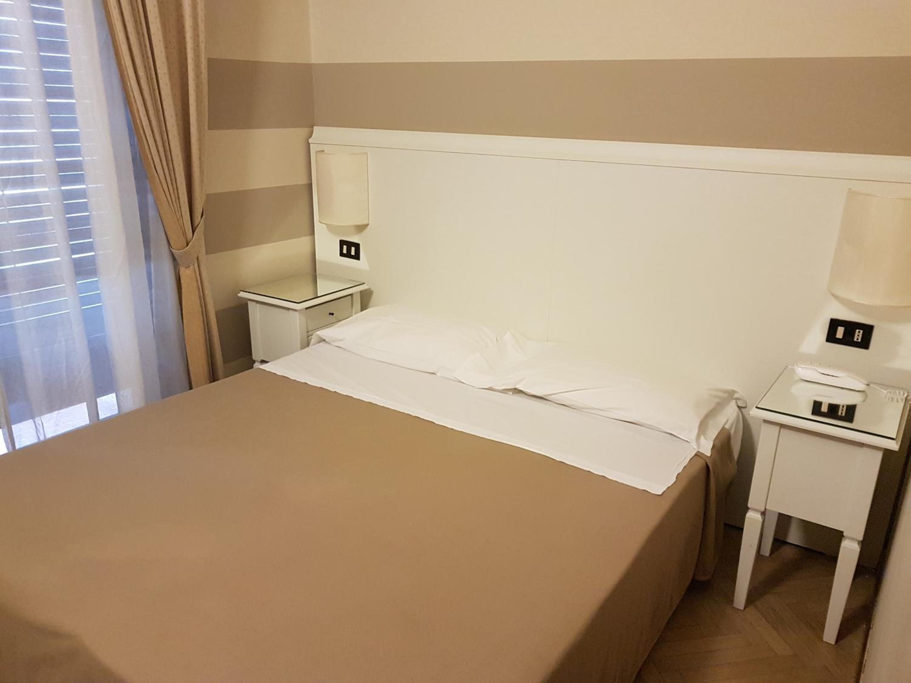 A hotel room with a single bed, two bedside tables, a nightstand, a lamp, and a window with curtains.