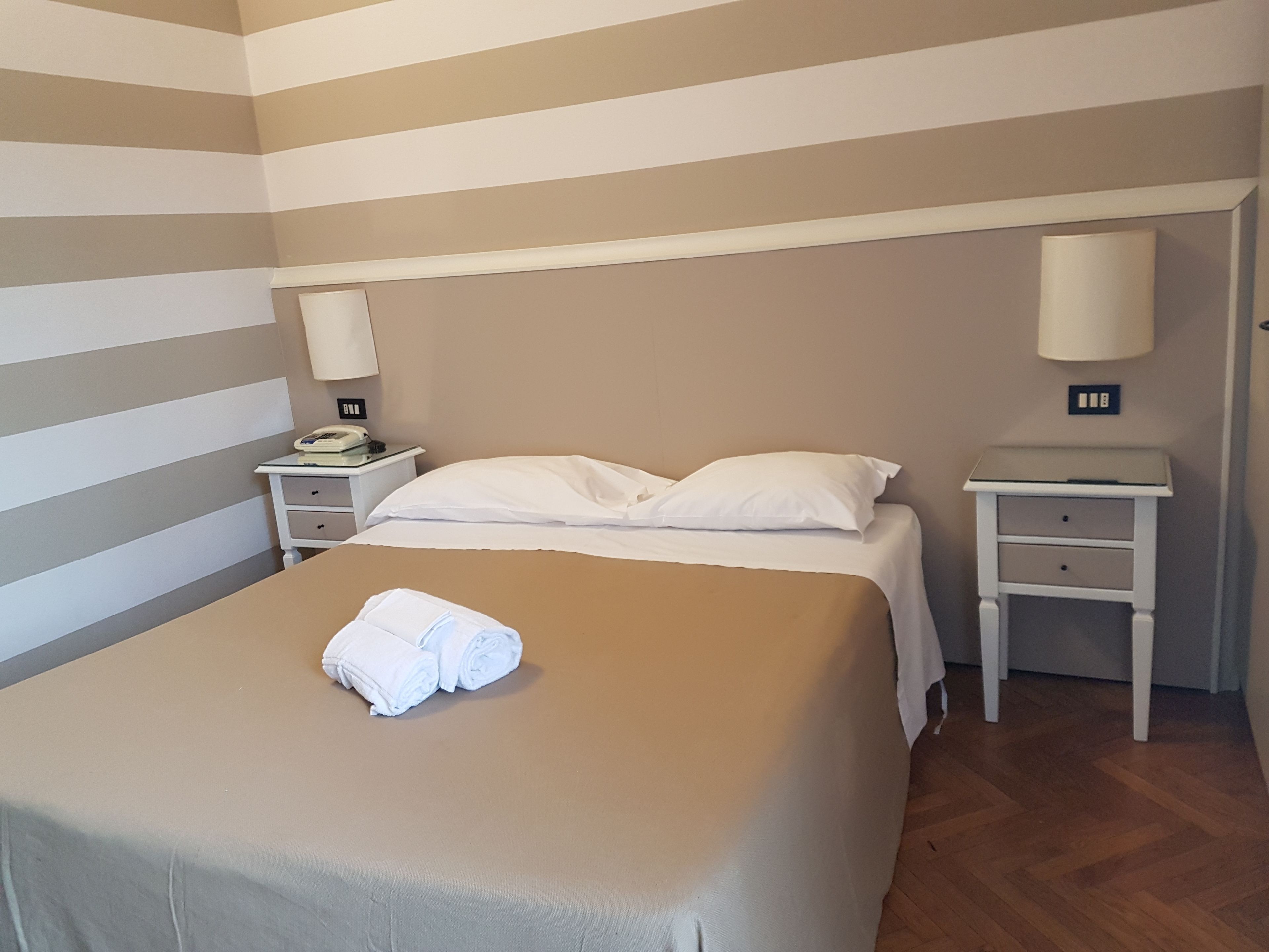 A hotel room with a bed, two bedside tables, a lamp on each table, and a folded towel on the bed.