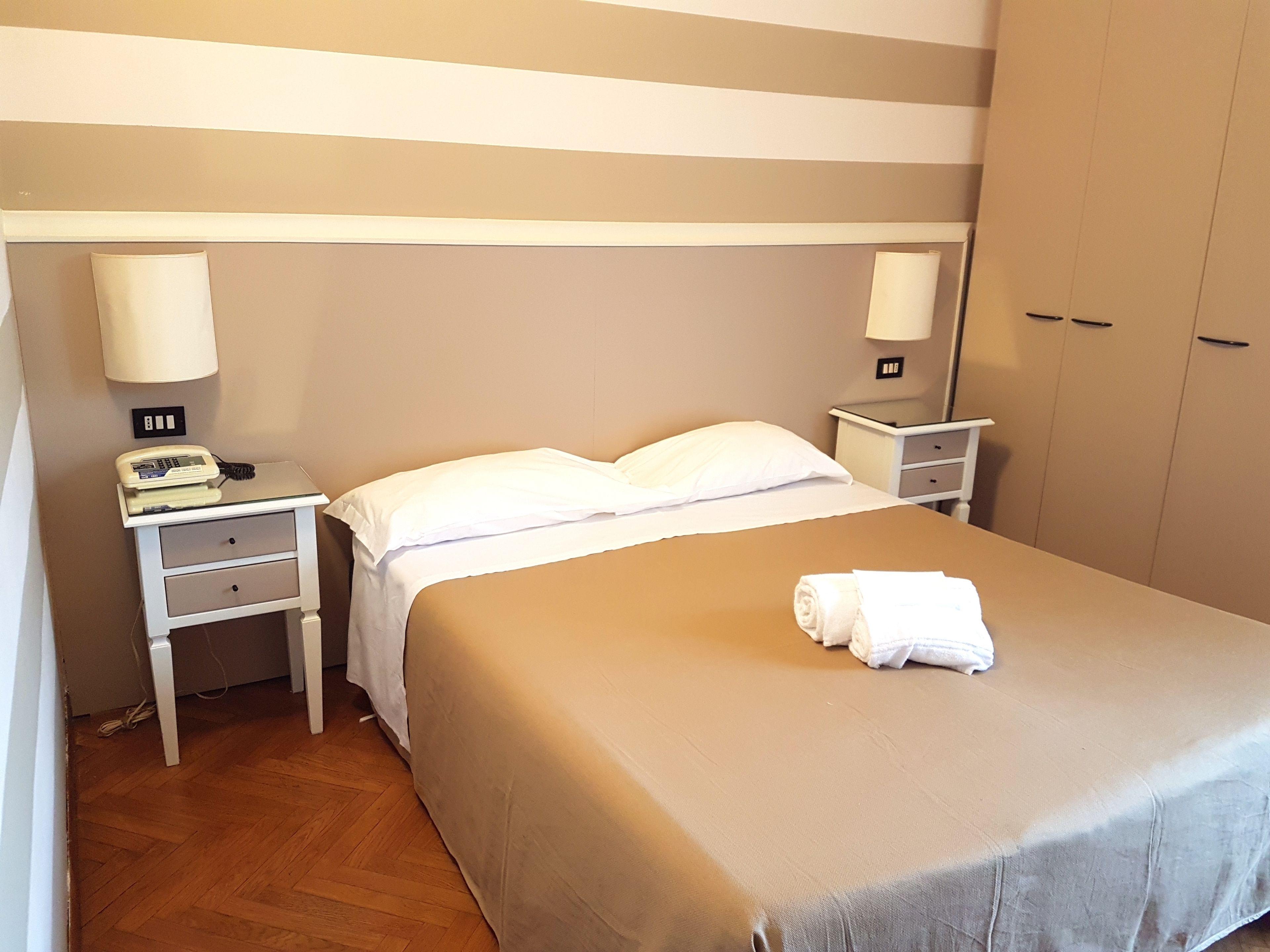 Una habitación de hotel con una cama, dos mesitas de noche, una lámpara, un teléfono y un armario.