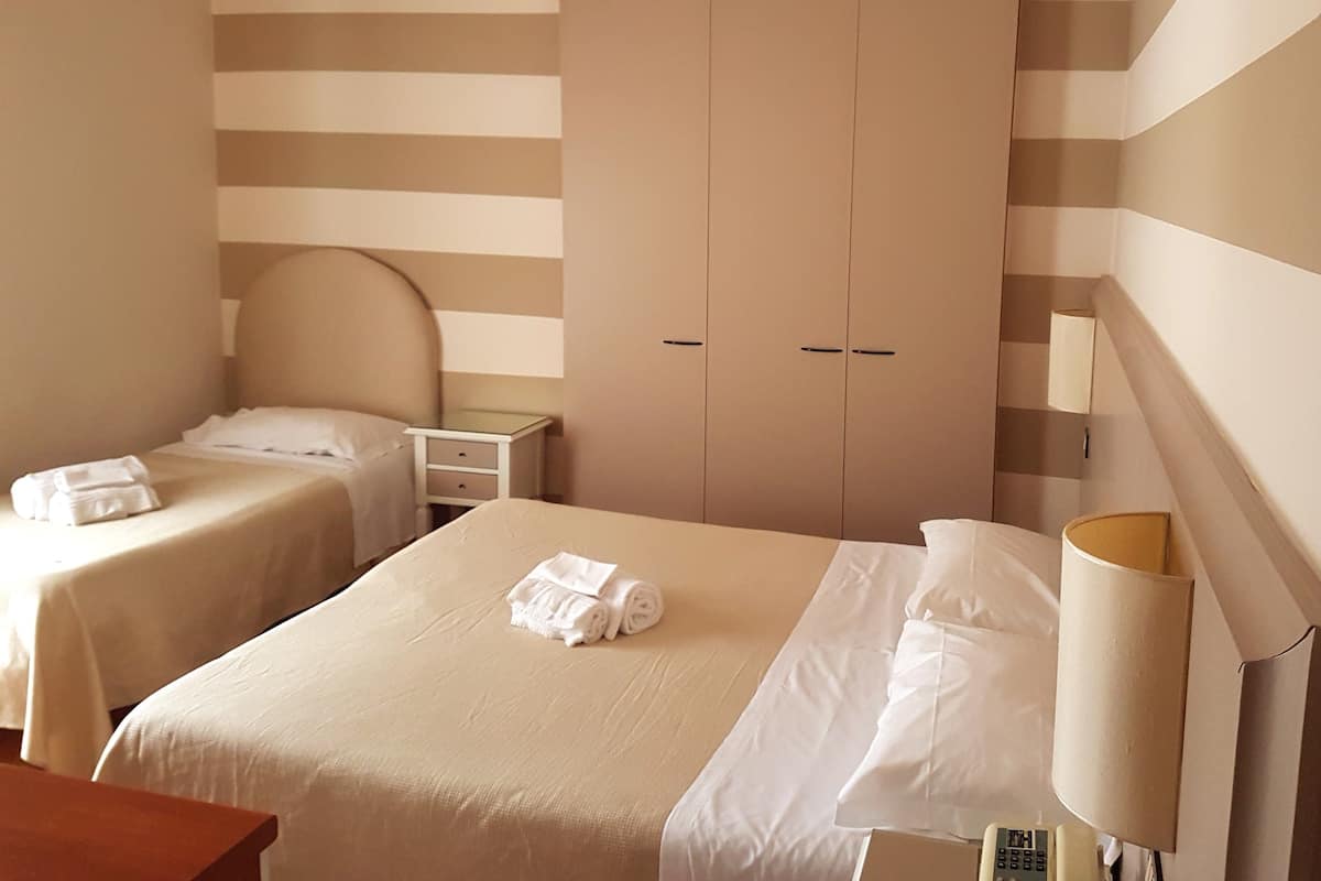 A hotel room with two beds, a wardrobe, a bedside table, a lamp, and an air conditioning unit.