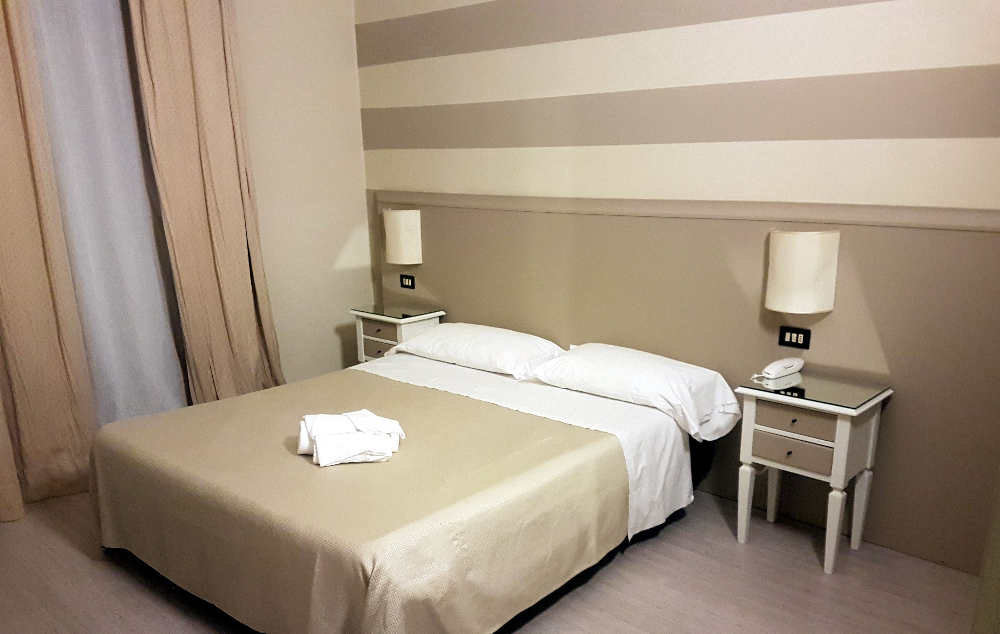 Habitación de hotel con una cama, dos mesitas de noche, una lámpara en cada una y un teléfono en una de ellas.